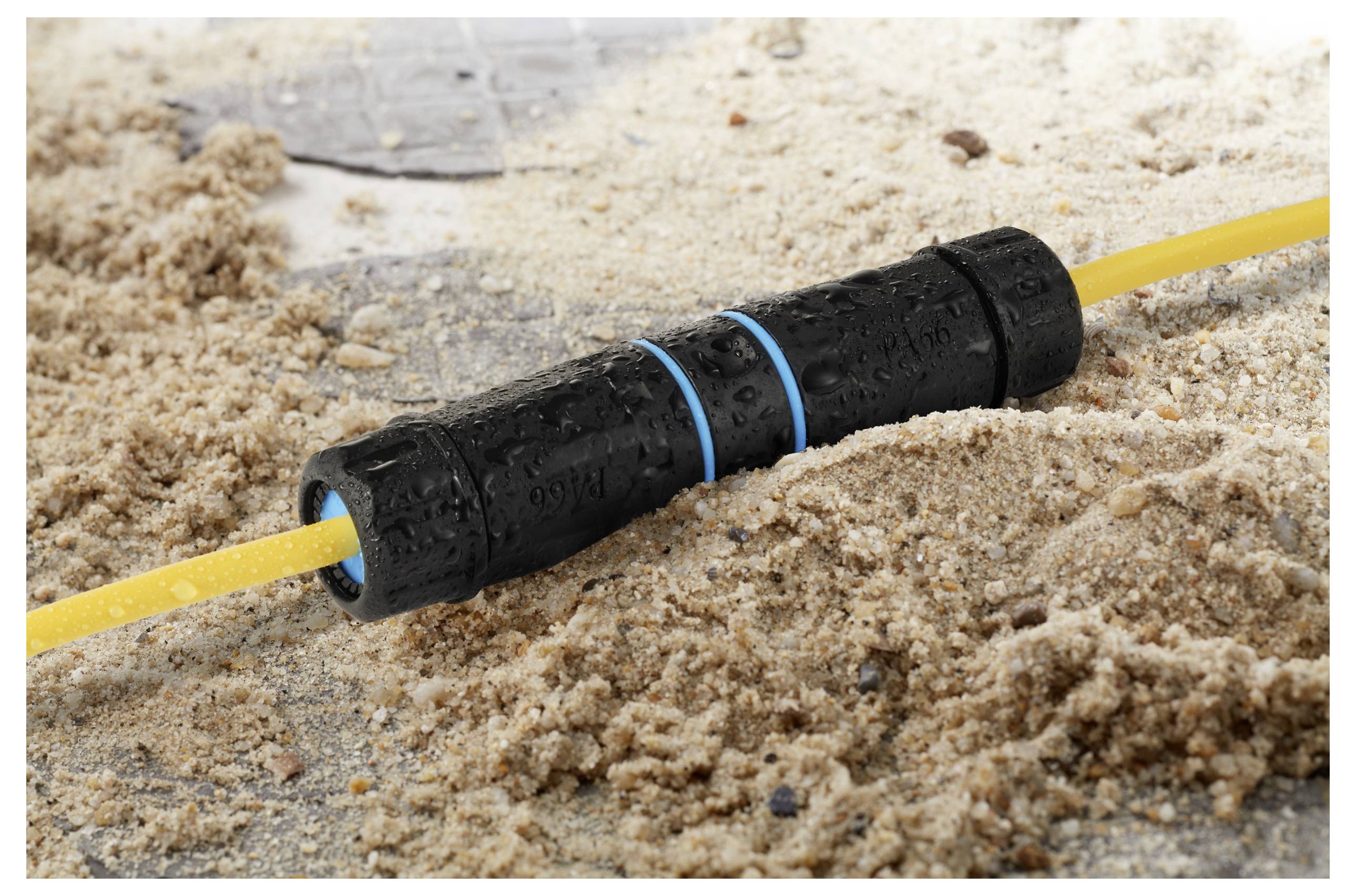 A black waterproof connector joins two yellow cables on sandy ground, indicating outdoor or rugged use.