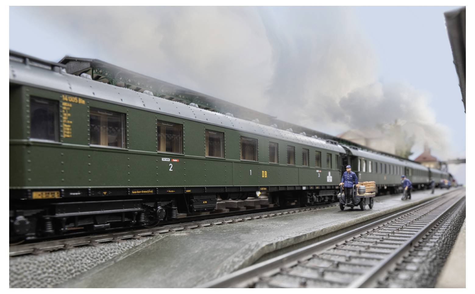 A model train in a station setting, featuring detailed green passenger cars, a platform, and a conductor with cargo on a cart.