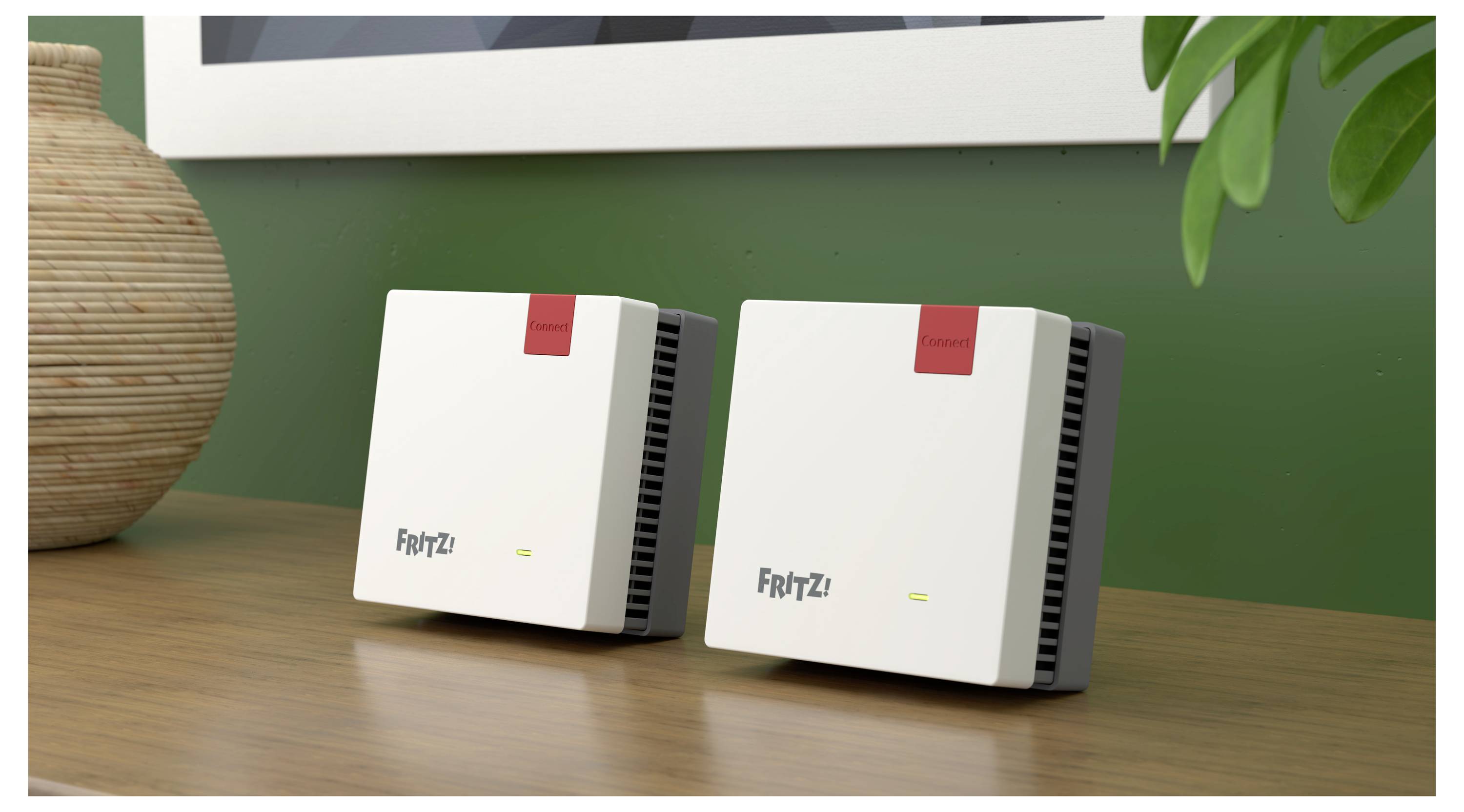 Two white FortiZi devices with red labels sit on a wooden table against a green wall, next to a plant and under a framed picture.