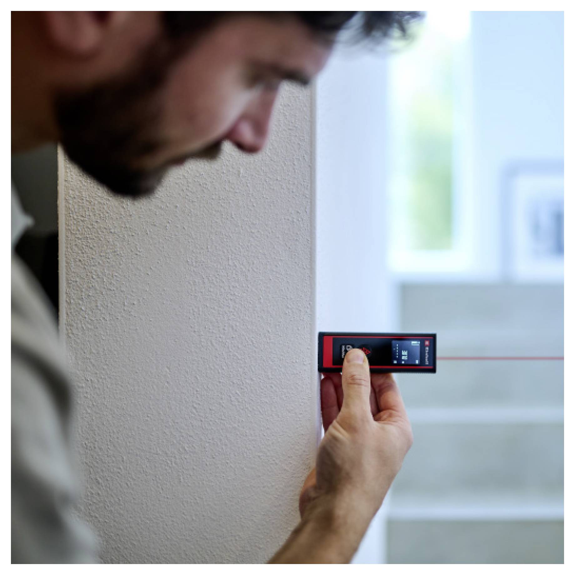 A person using a digital laser measuring tool against a wall with stairs visible in the background.
