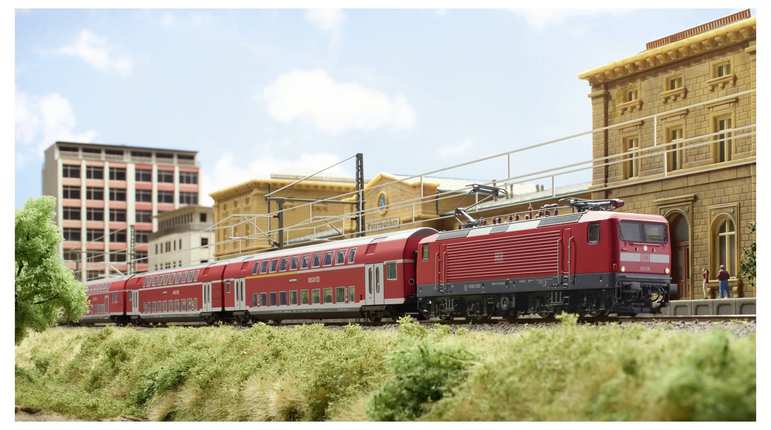 A red train passes through a model city with detailed buildings and greenery, showcasing a railway scene with urban architecture.
