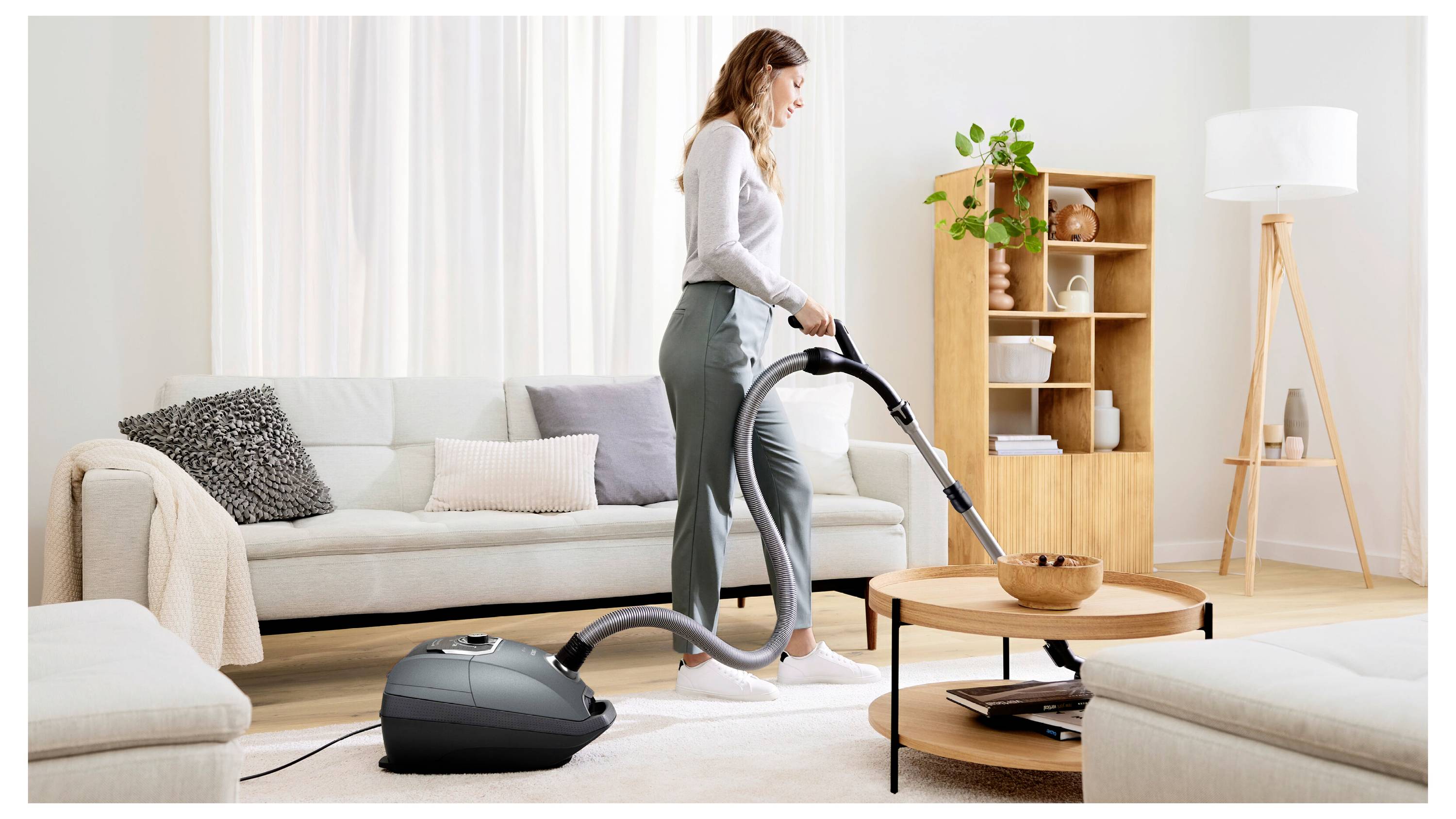 A person vacuuming a clean, modern living room with light decor, including a sofa, coffee table, and shelves with plants and books.