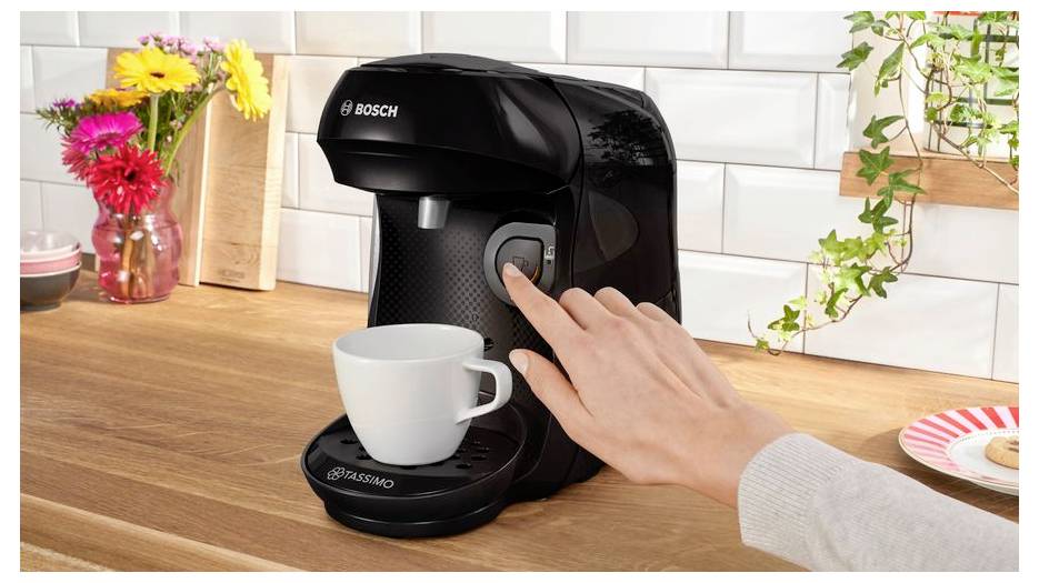 A hand operates a black coffee machine with a white mug beneath. Bright flowers and a plant decorate the wooden countertop.