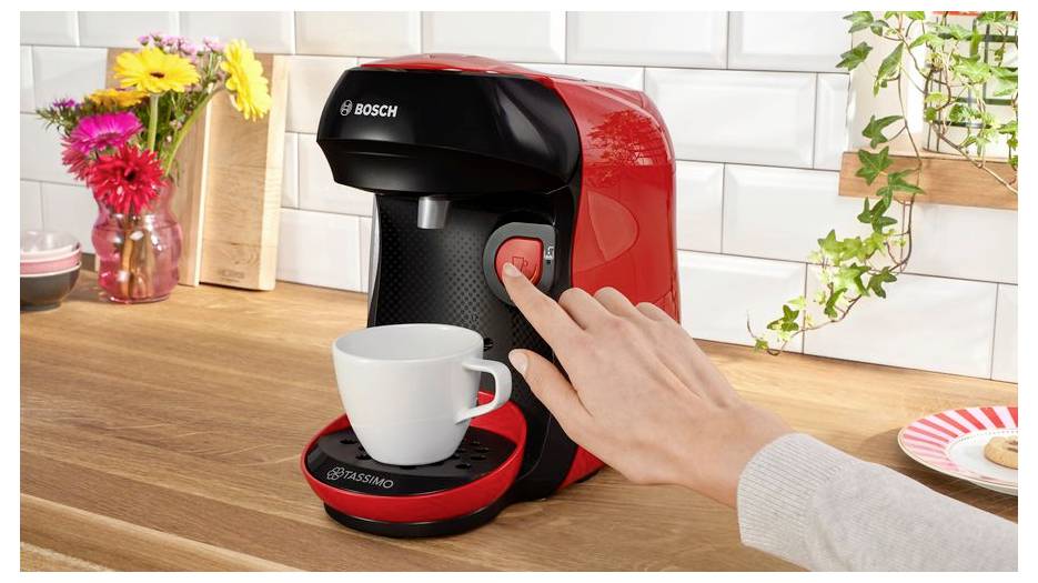 A person pressing a button on a red coffee machine to make coffee, with a white cup placed on the drip tray and flowers in the background.