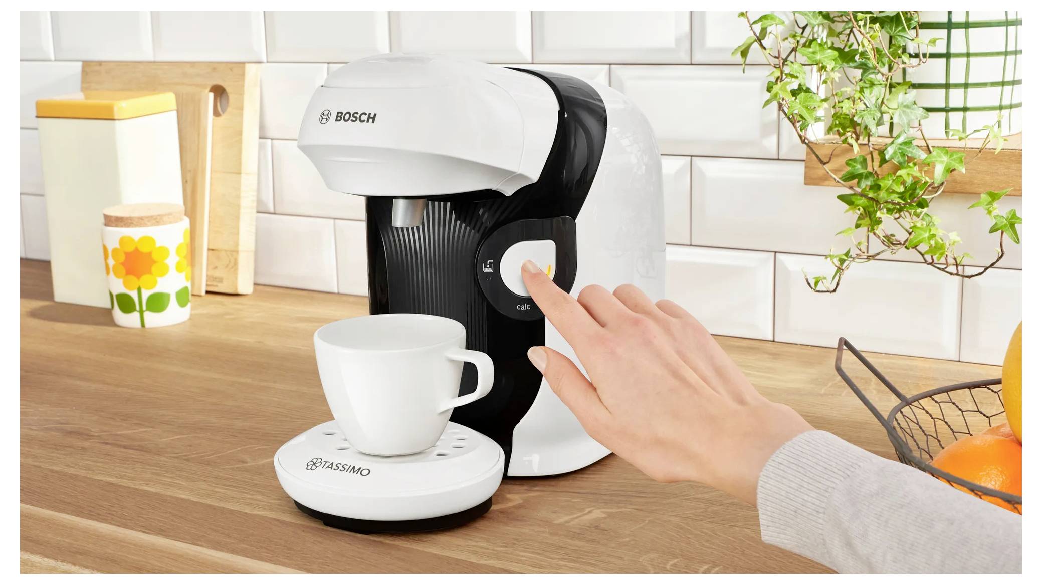 A person presses a button on a single-serve coffee maker, placed on a kitchen counter with a mug ready underneath.