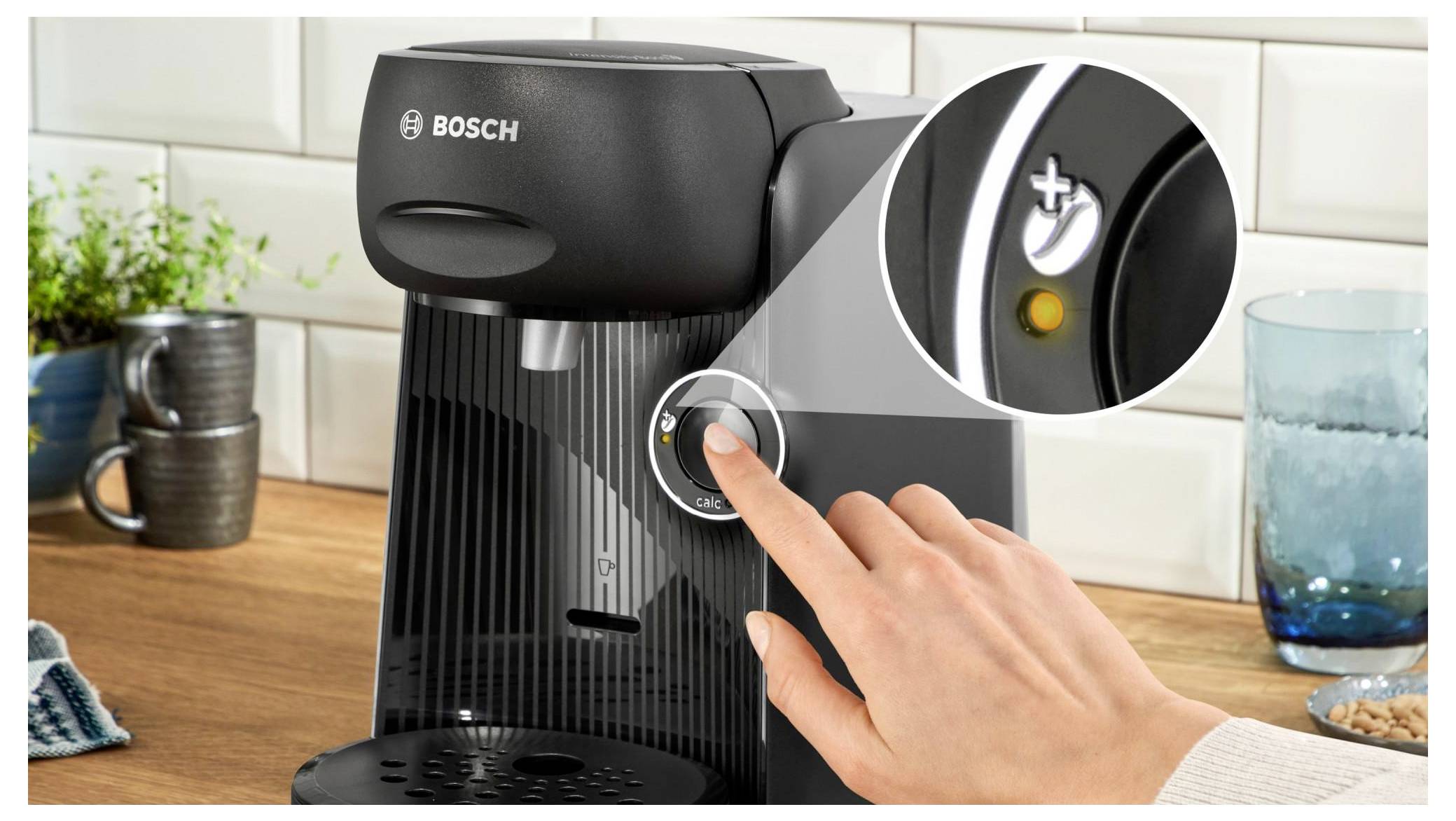 A hand presses a button on a Bosch coffee maker with a focus on the control panel. The kitchen setting includes cups and dishes nearby.