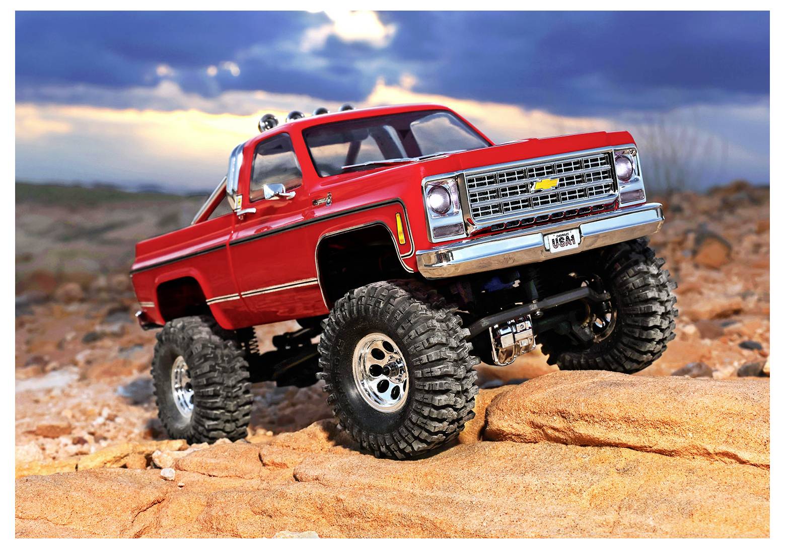 A lifted red toy truck with large tires on rocky terrain against a dramatic sky.