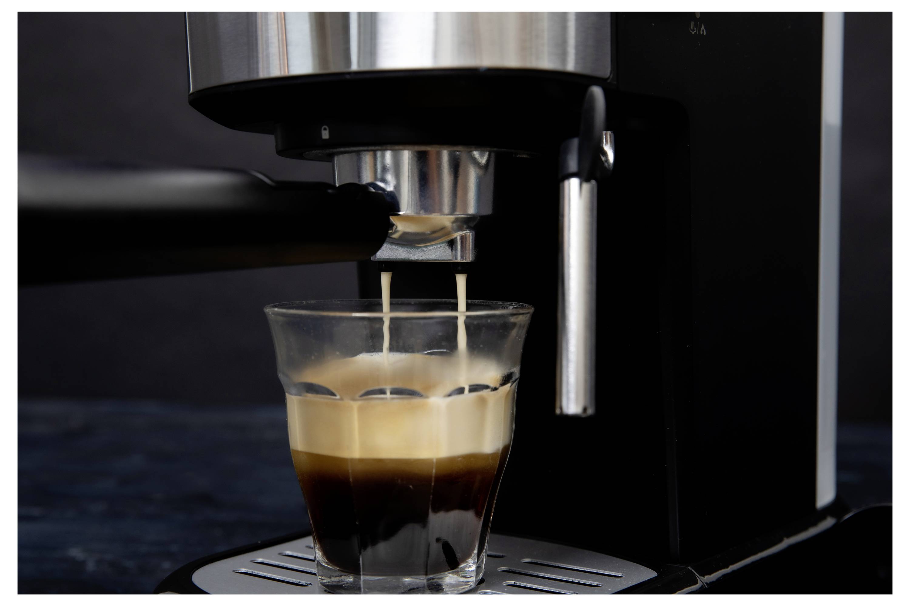 An espresso machine pours steaming coffee into a clear glass, forming layers of crema and dark liquid on a dark background.