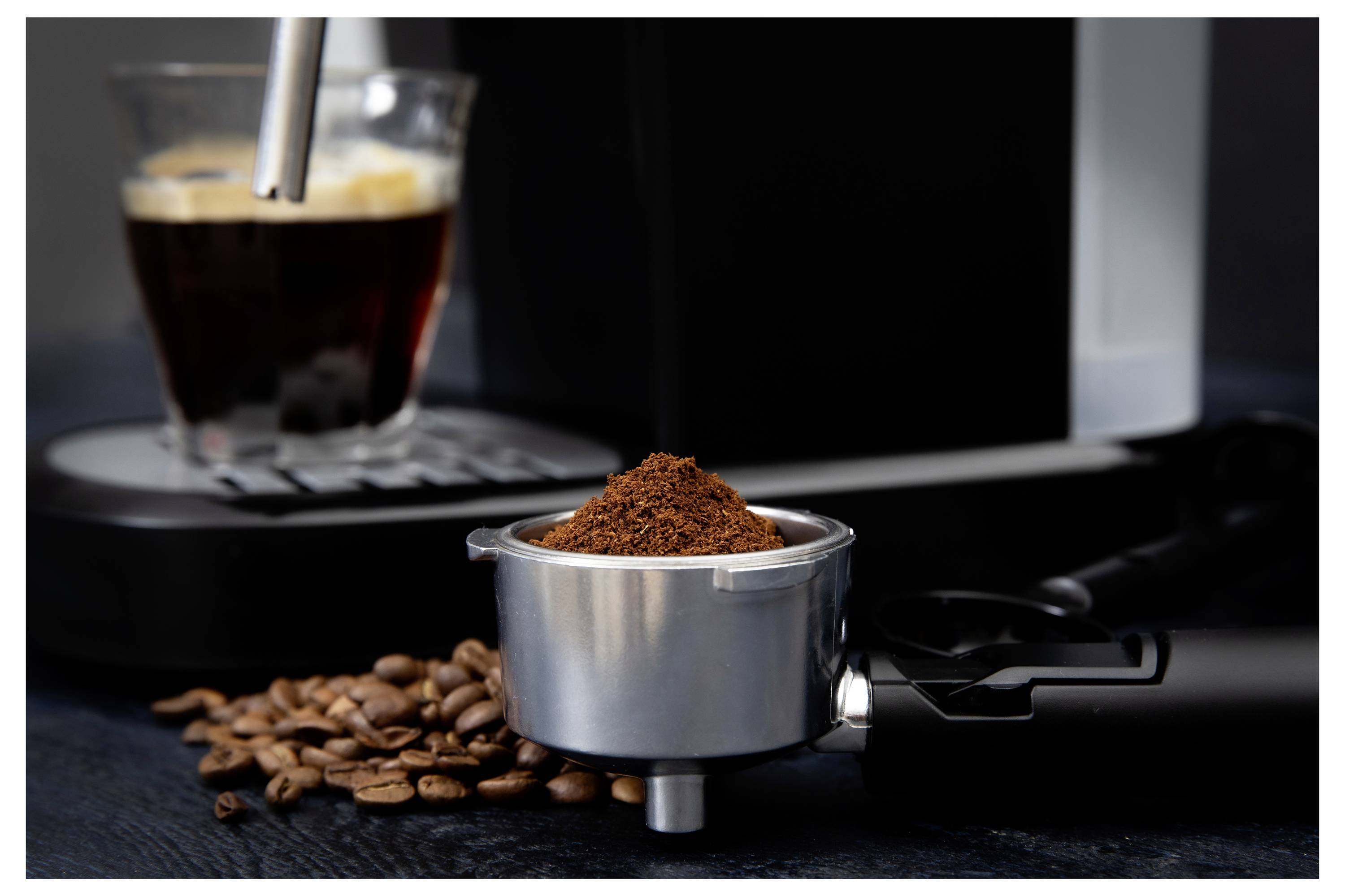 A portafilter filled with ground coffee is placed next to scattered coffee beans and an espresso machine, with a brewed espresso in the background.