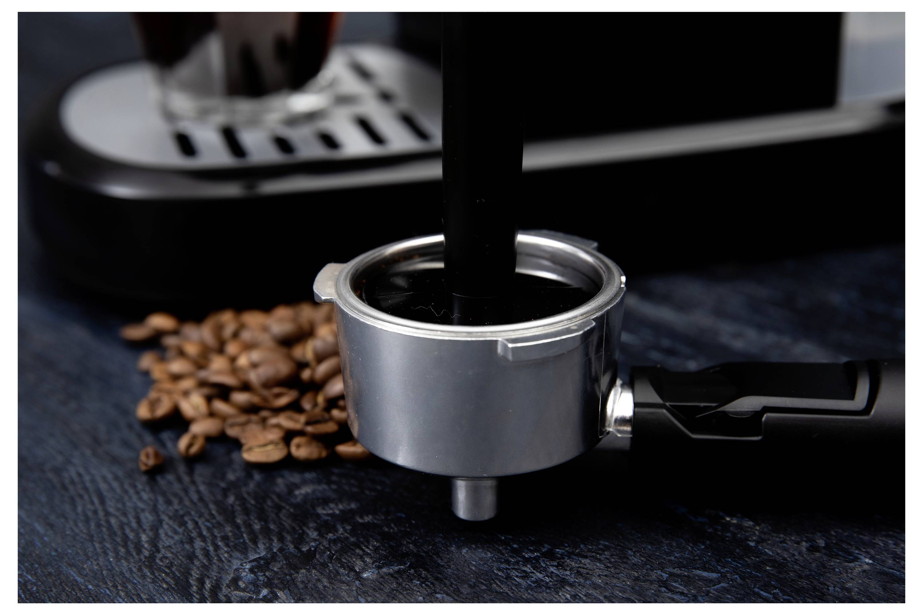 A portafilter is being filled with ground coffee next to scattered coffee beans. An espresso machine is partially visible in the background.