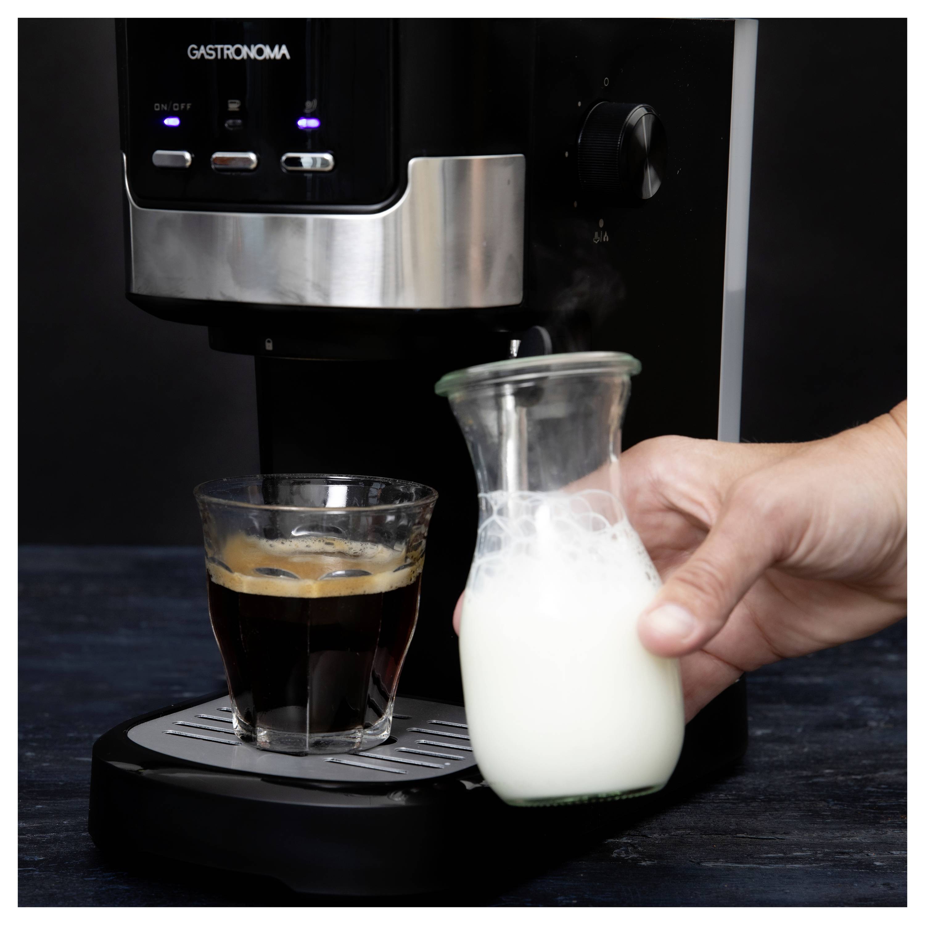 A black coffee machine is dispensing coffee into a glass. A hand holds a small glass jug filled with milk beside the machine.