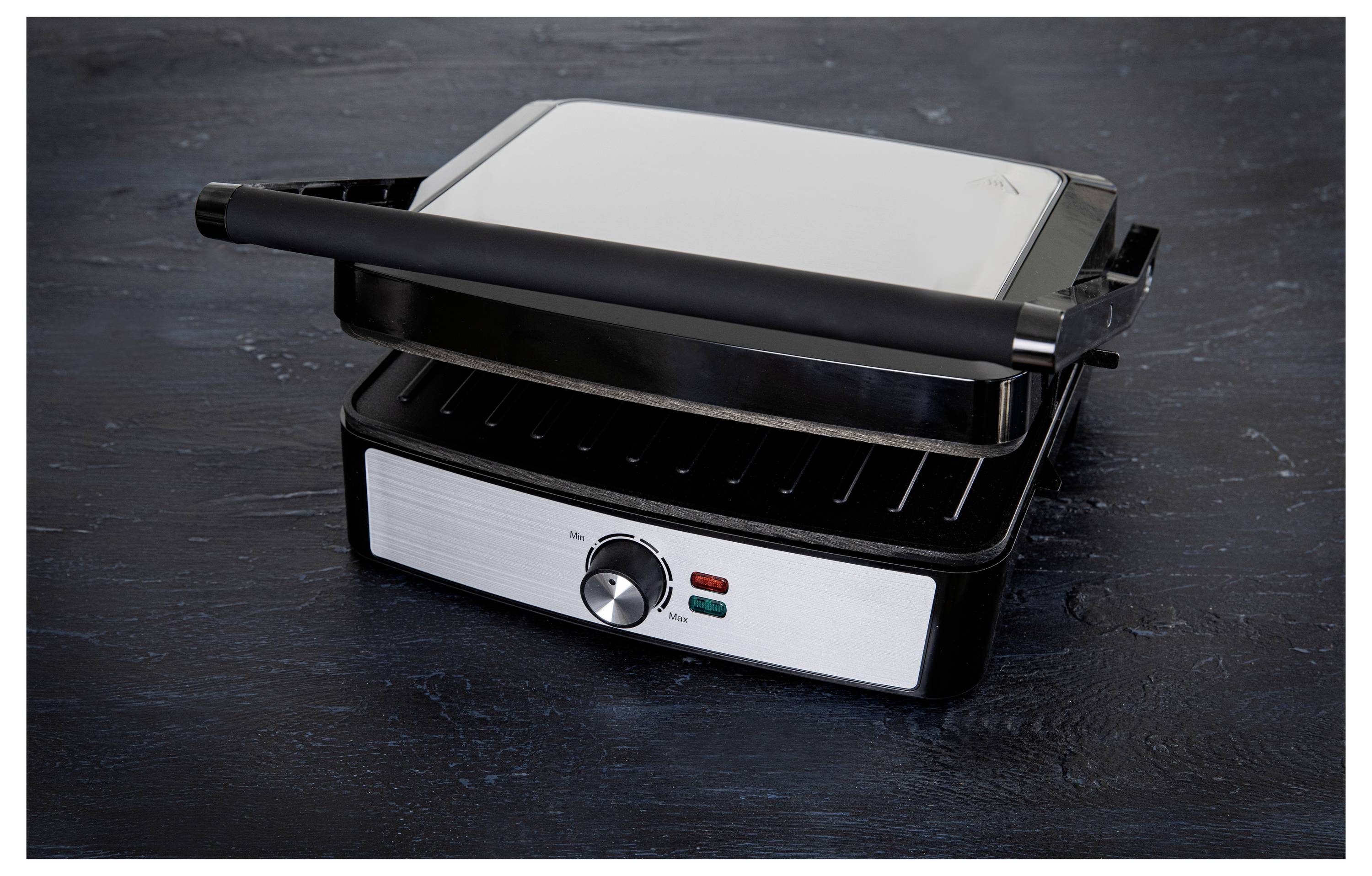 A silver and black electric grill with a handle sits on a dark surface. The grill has an adjustable temperature dial and indicator lights.