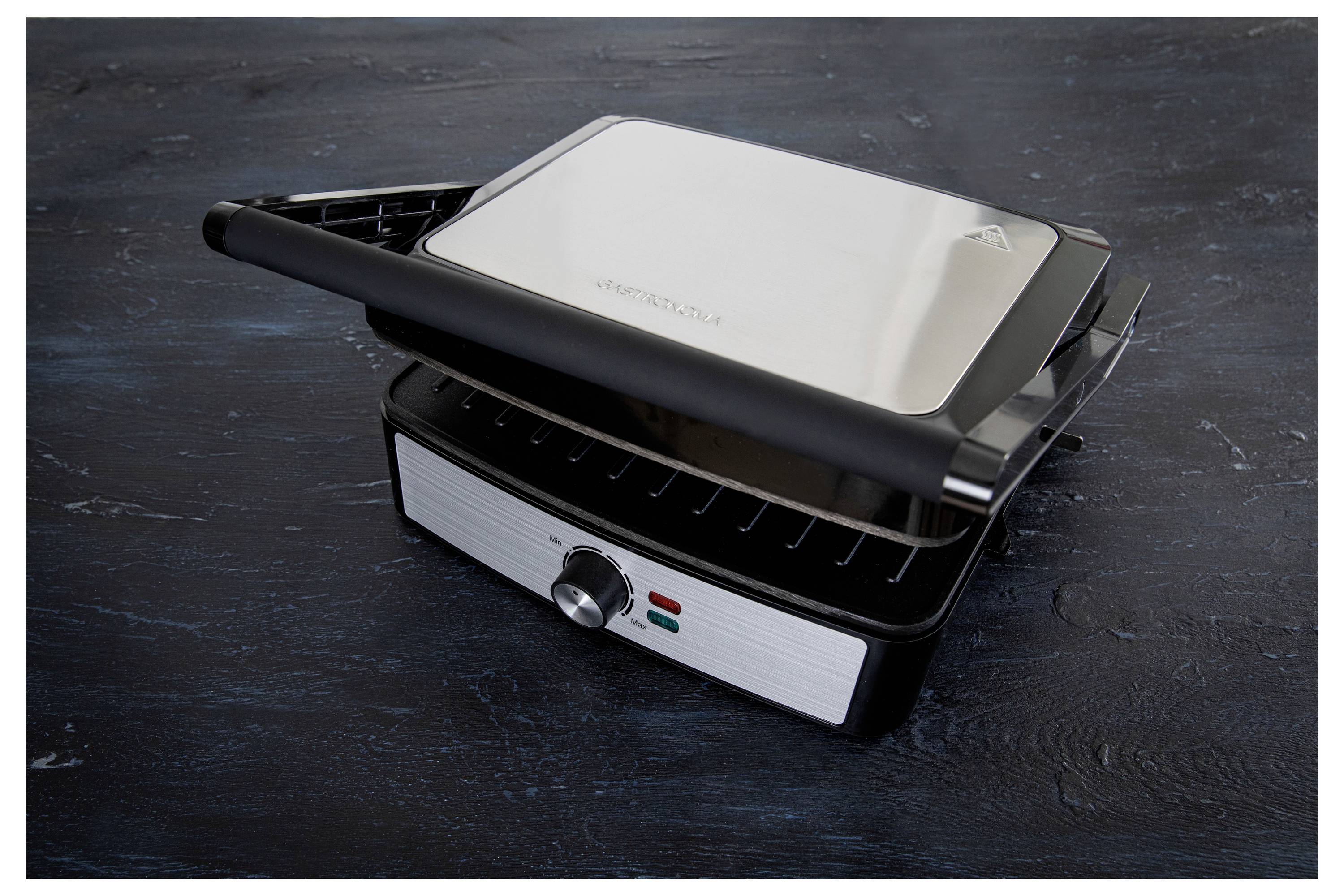 A stainless steel panini press with a black handle on a dark surface. The knob on the front features two indicator lights.