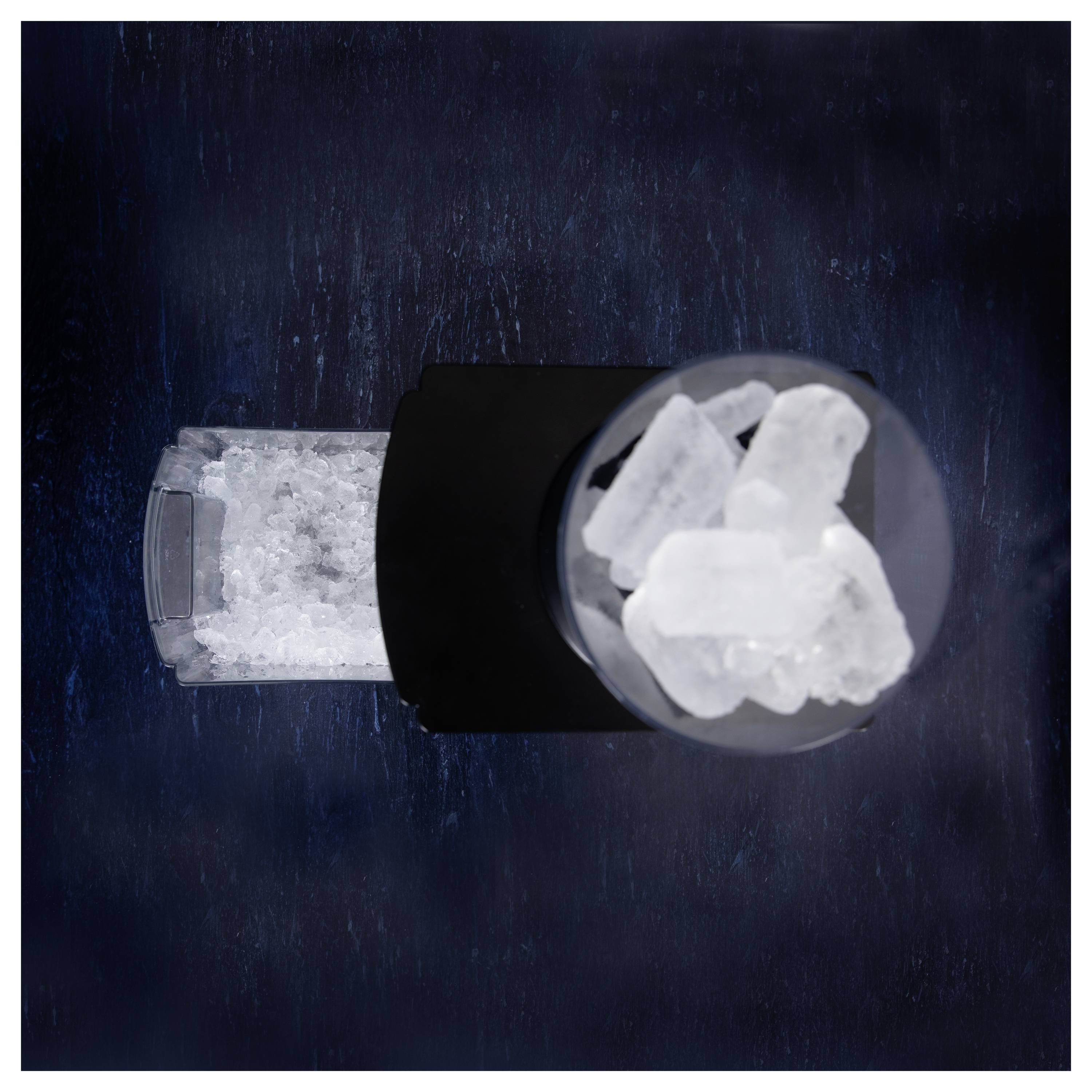 A top-down view of a black manual ice grinder. Large ice cubes are visible in the top compartment, with crushed ice in the side container.