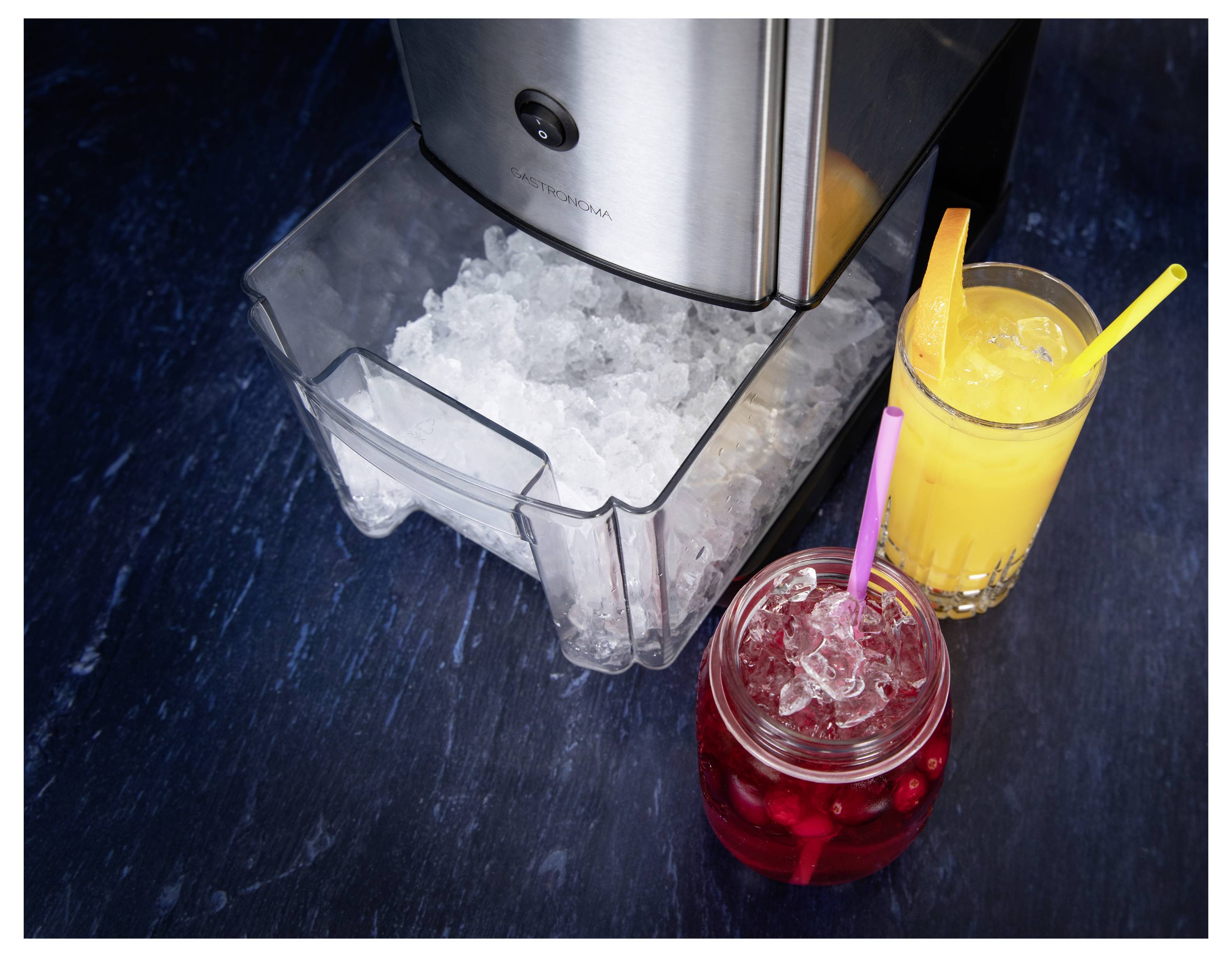 Ice machine with open drawer full of ice next to two drinks with straws, one red with berries and one yellow on a dark blue surface.