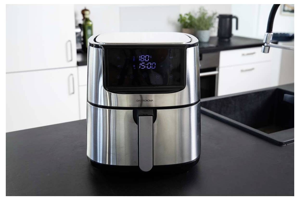 A stainless steel air fryer on a kitchen counter with digital display showing '180°C'. Kitchenware and plants are in the background.