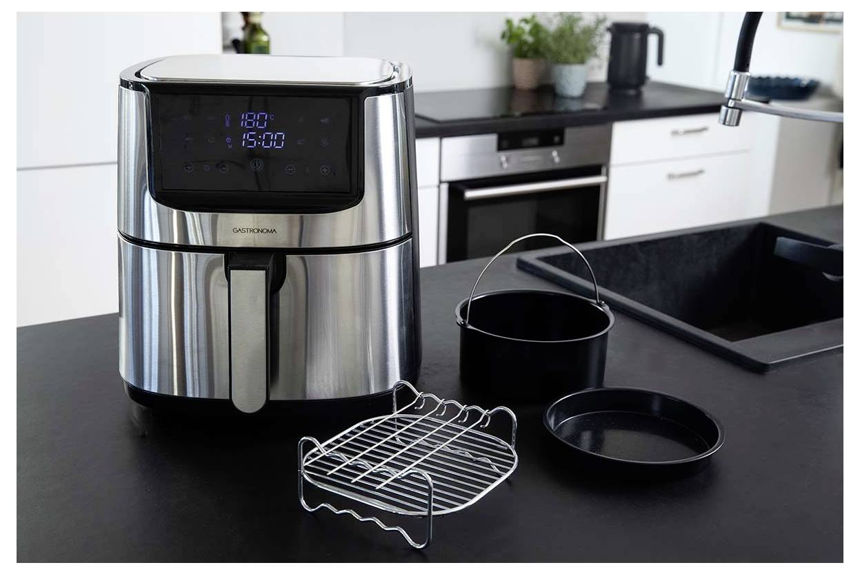 A stainless steel air fryer on a kitchen counter with a digital display showing 180°C and 15:00. Nearby are cooking accessories.