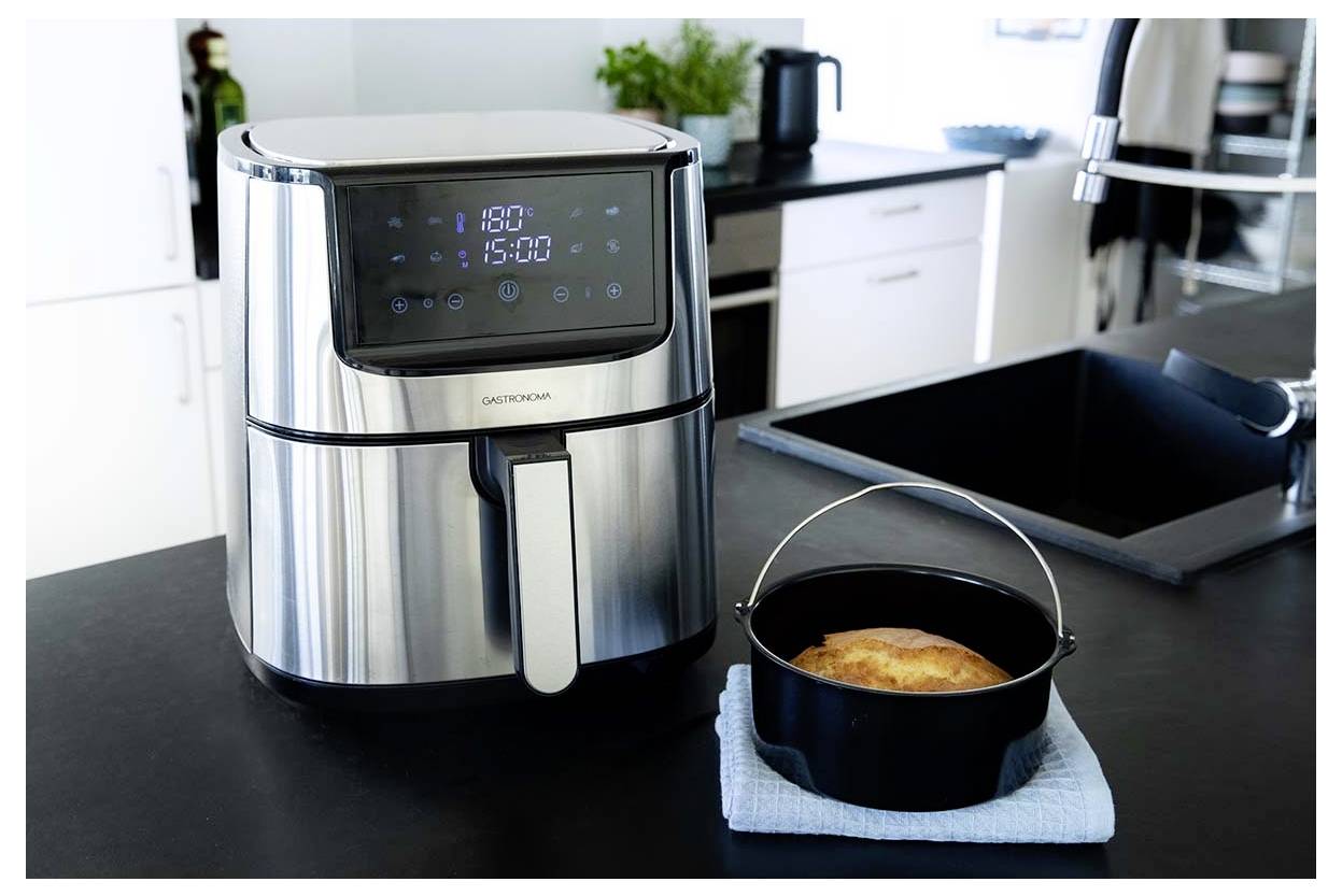 A stainless steel air fryer on a kitchen counter displaying '180°C' on its screen. A cooked dish sits beside it on a blue cloth.