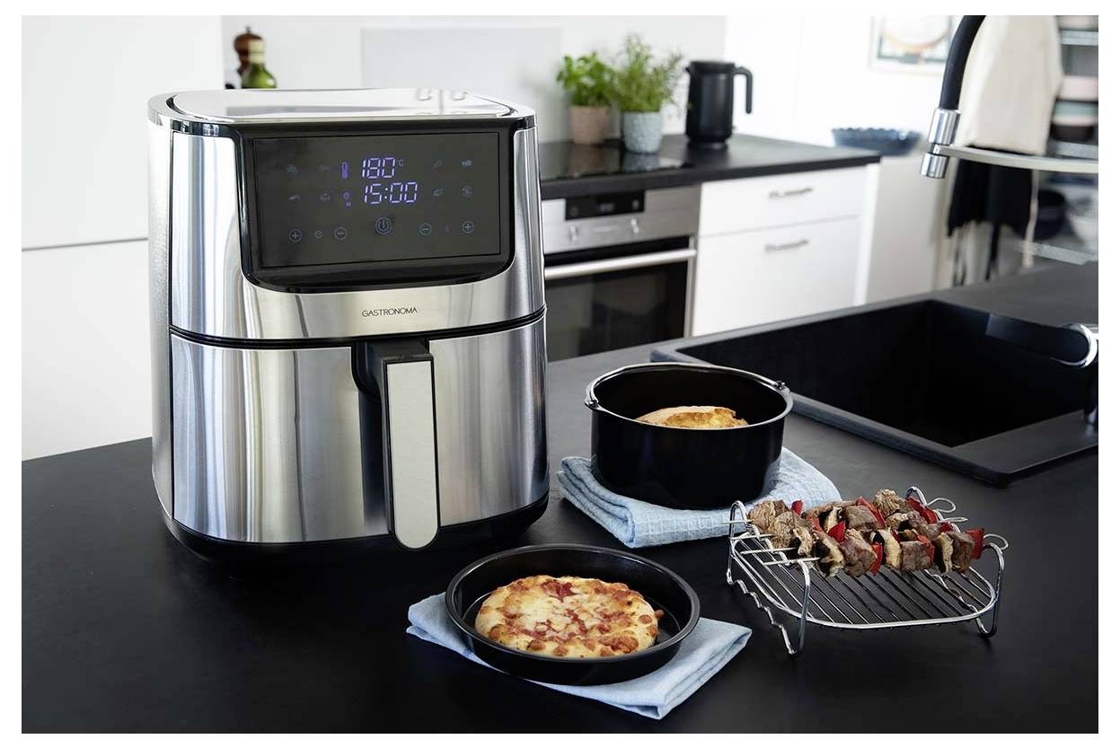 A modern kitchen with a stainless steel air fryer displaying '180°' and '15:00'. Nearby, a pizza, a casserole, and skewers are prepared.