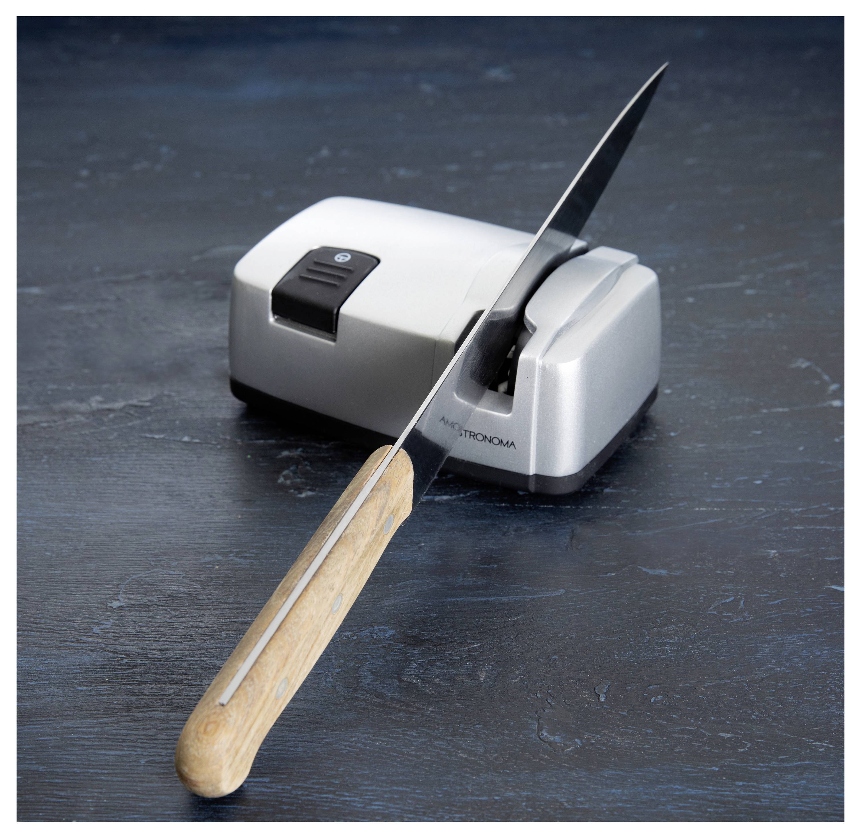 A sharp kitchen knife with a wooden handle rests on an electric knife sharpener, set against a dark surface.