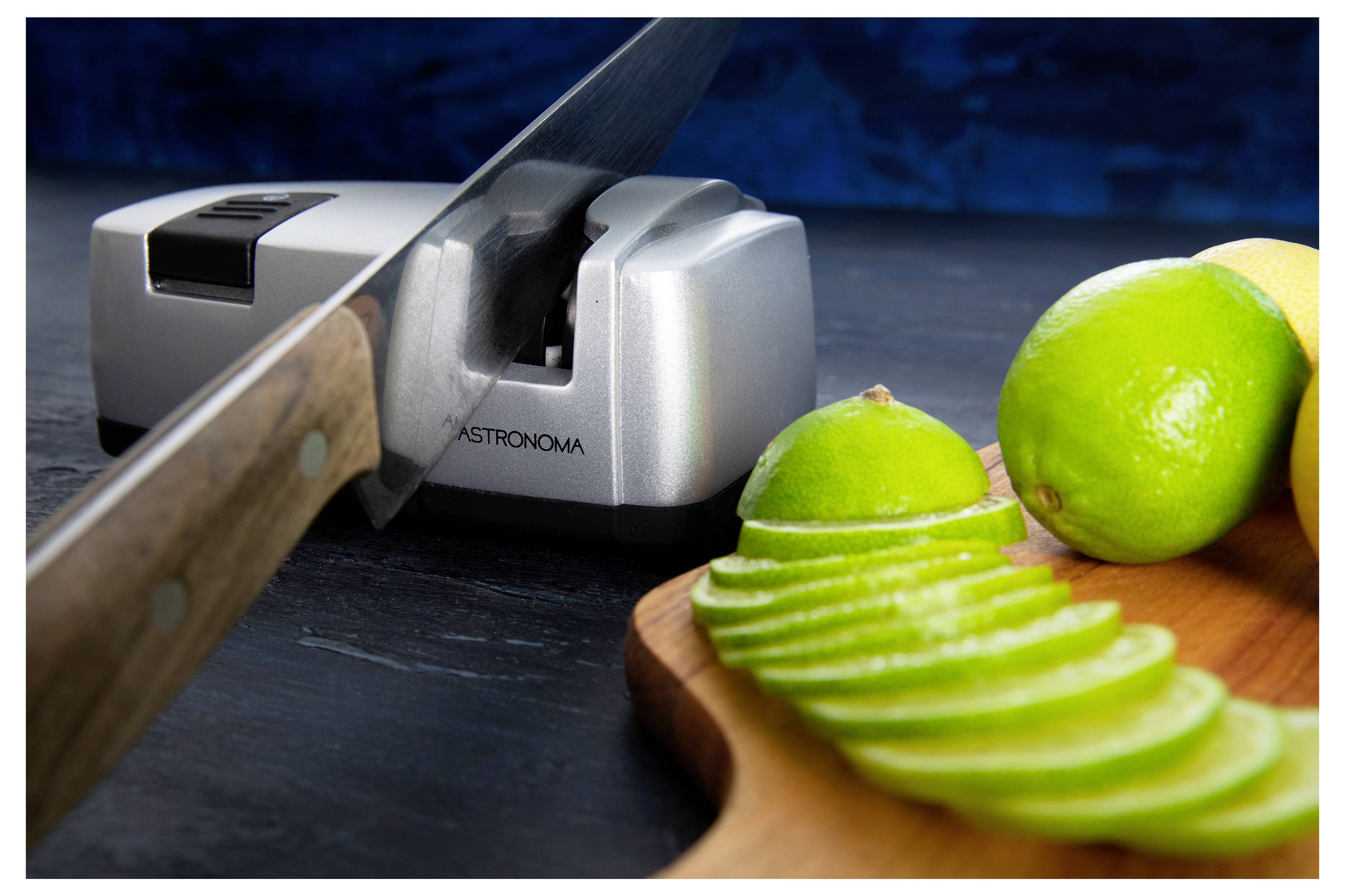 A knife blade rests on an electric sharpener. In the foreground, sliced limes on a wooden board are visible, indicating kitchen preparation.