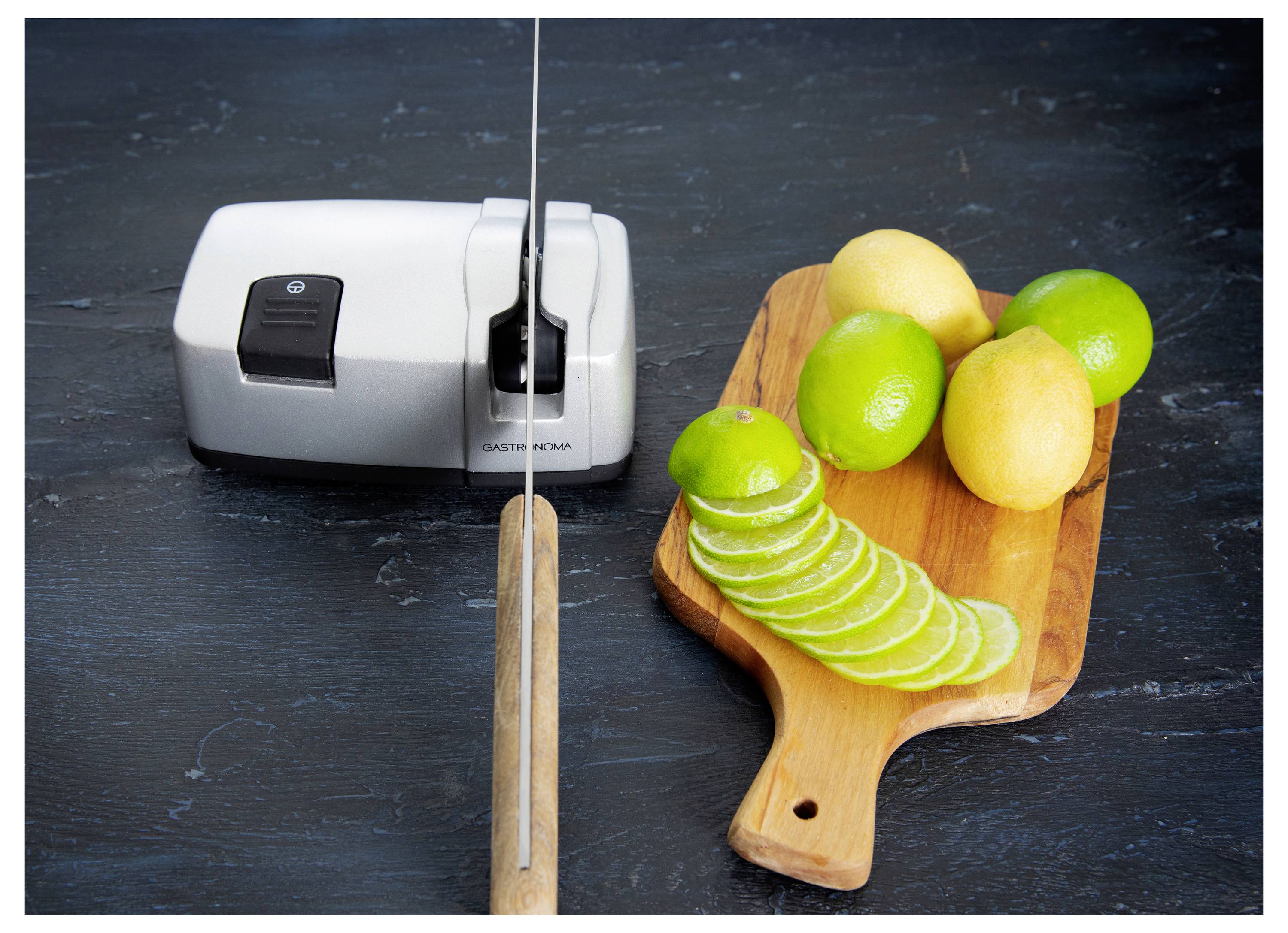 A knife sharpener next to a cutting board with sliced limes and whole lemons and limes on a dark countertop.