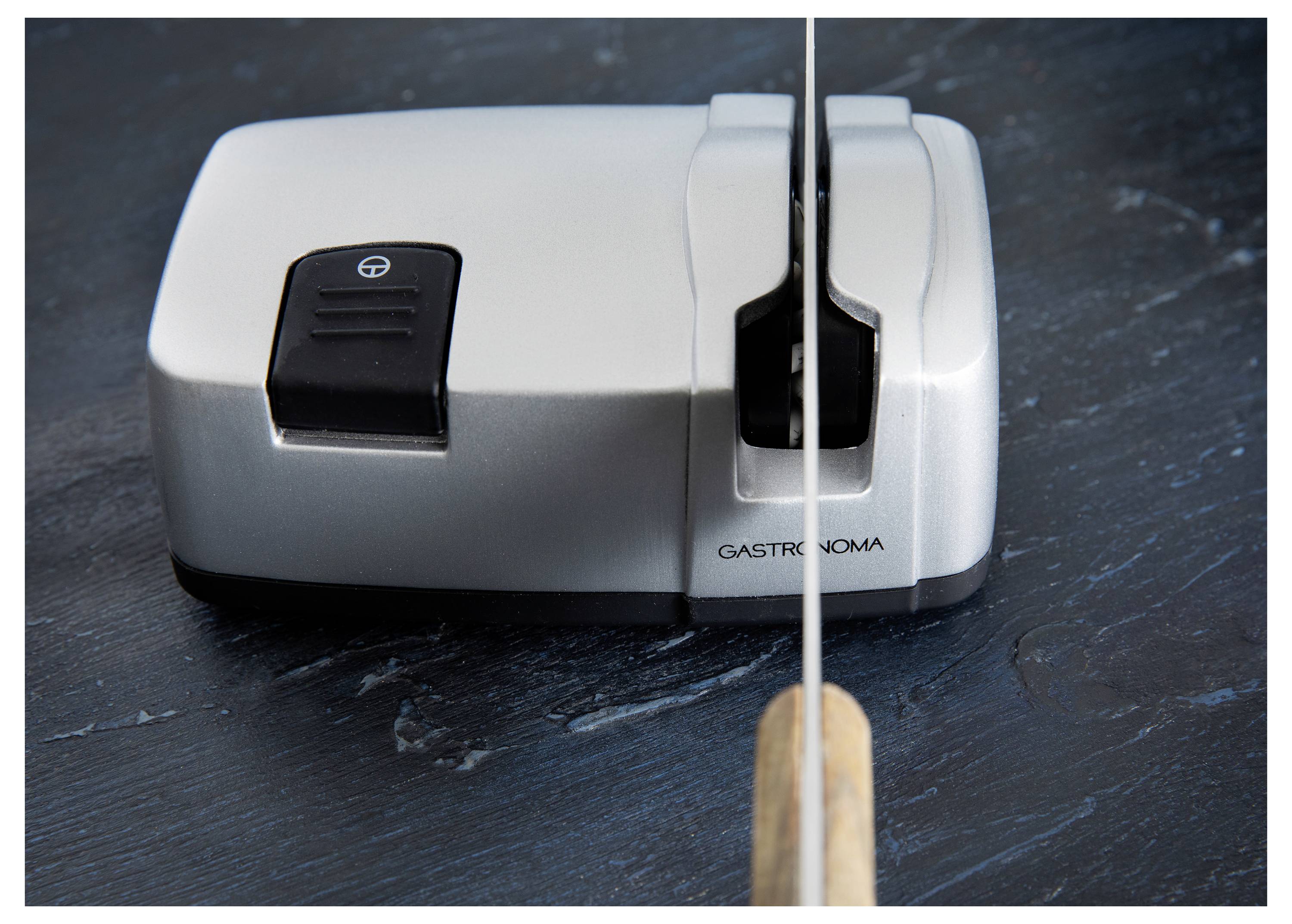 A knife being sharpened on a silver electric sharpener placed on a dark surface.