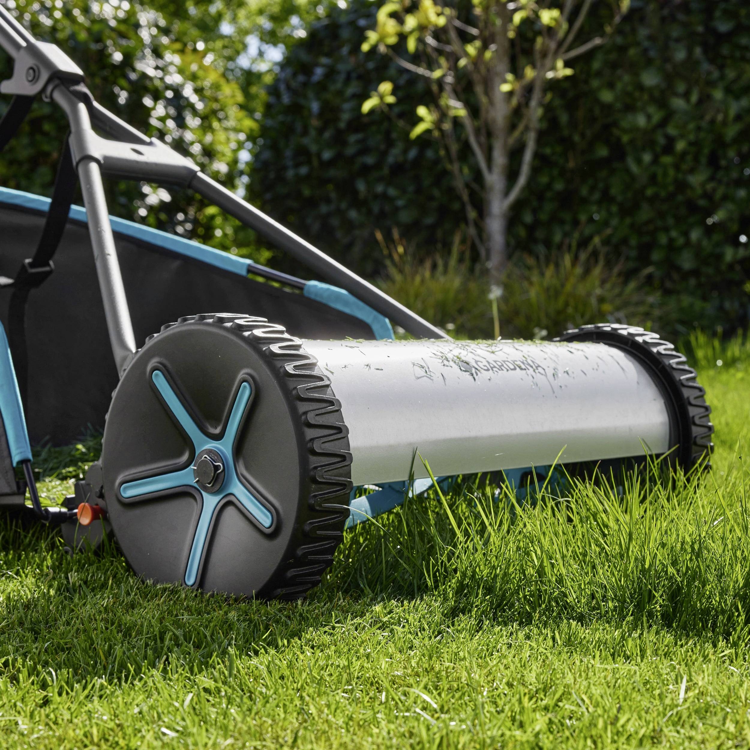 A close-up shot of a manual push lawn mower on green grass with a silver cylindrical drum and black wheels in a garden setting.
