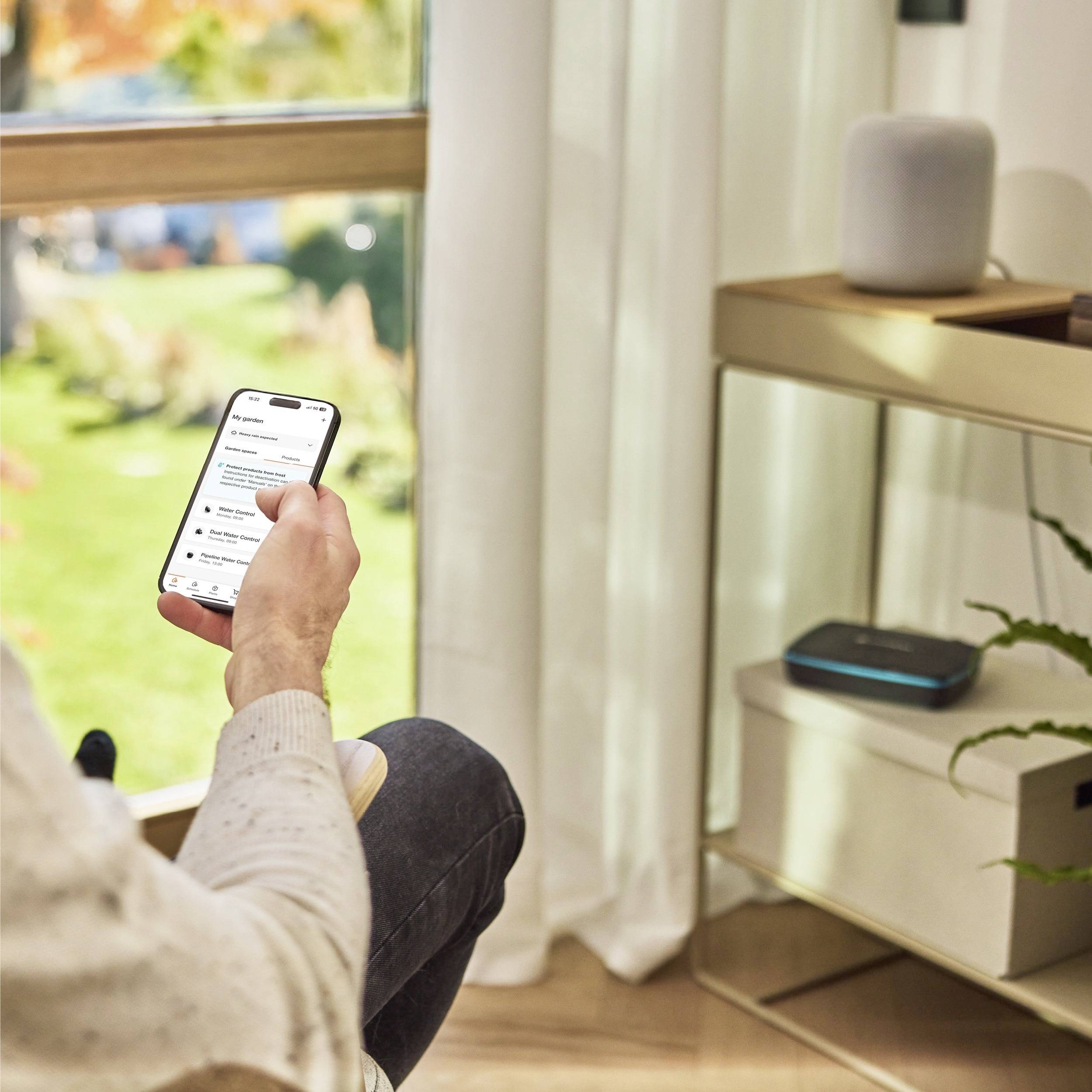 A person sits indoors holding a smartphone, viewing notifications. Nearby are a smart speaker and a router on a shelf.