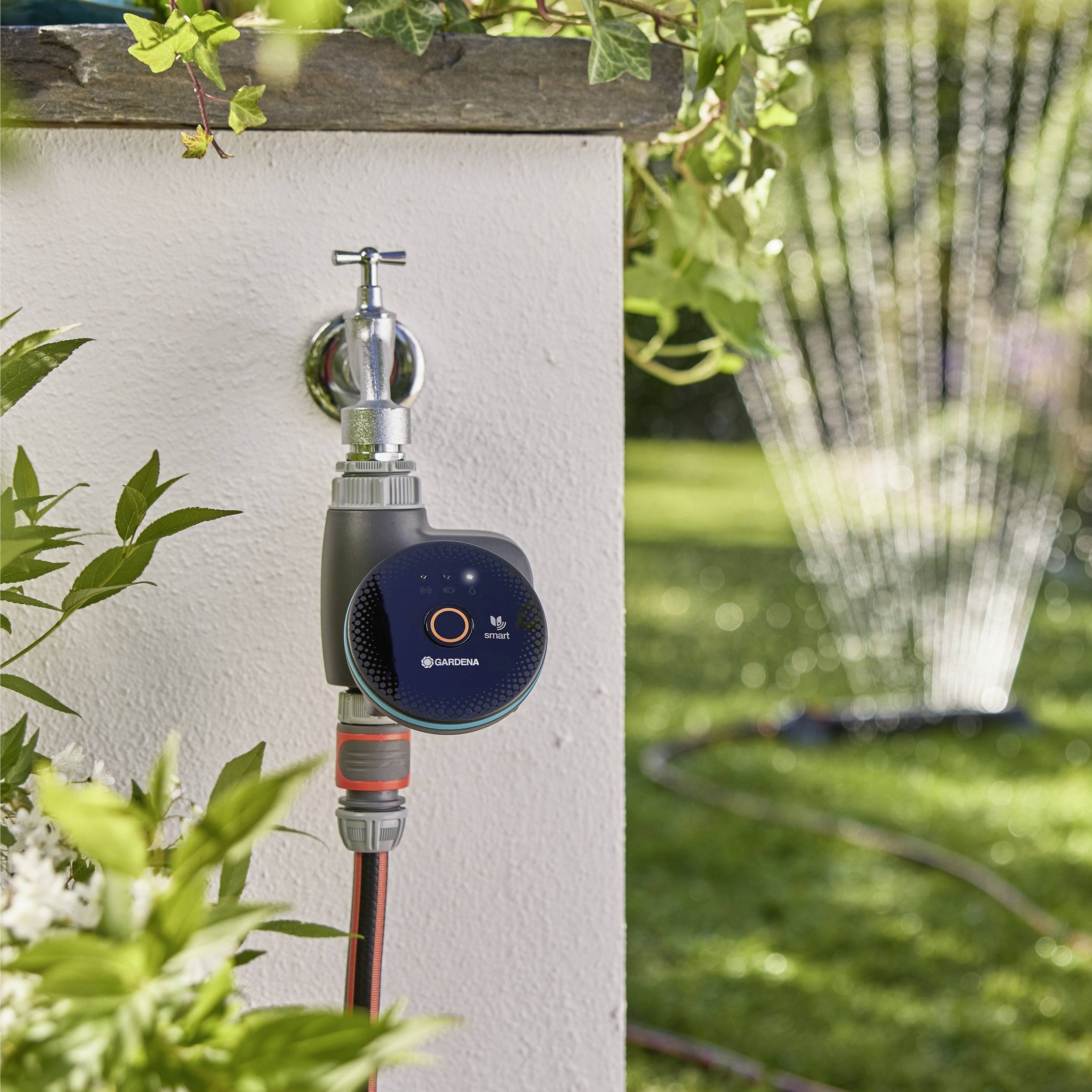 A garden wall-mounted water timer controls a hose, set in a sunny garden with greenery. A sprinkler waters the grass in the background.