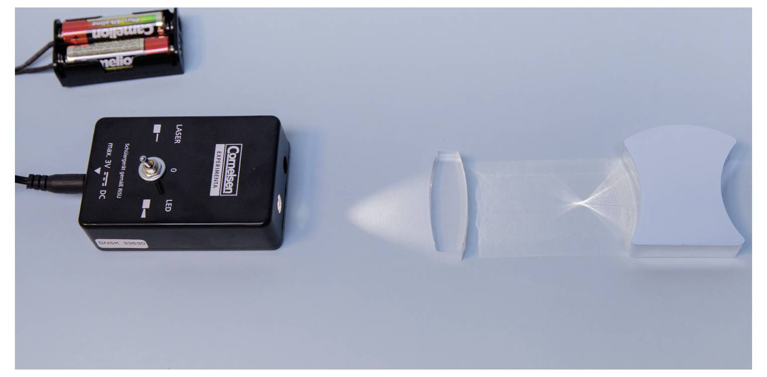 A black laser module projects a light beam towards a concave mirror on a blue surface, demonstrating light reflection principles.