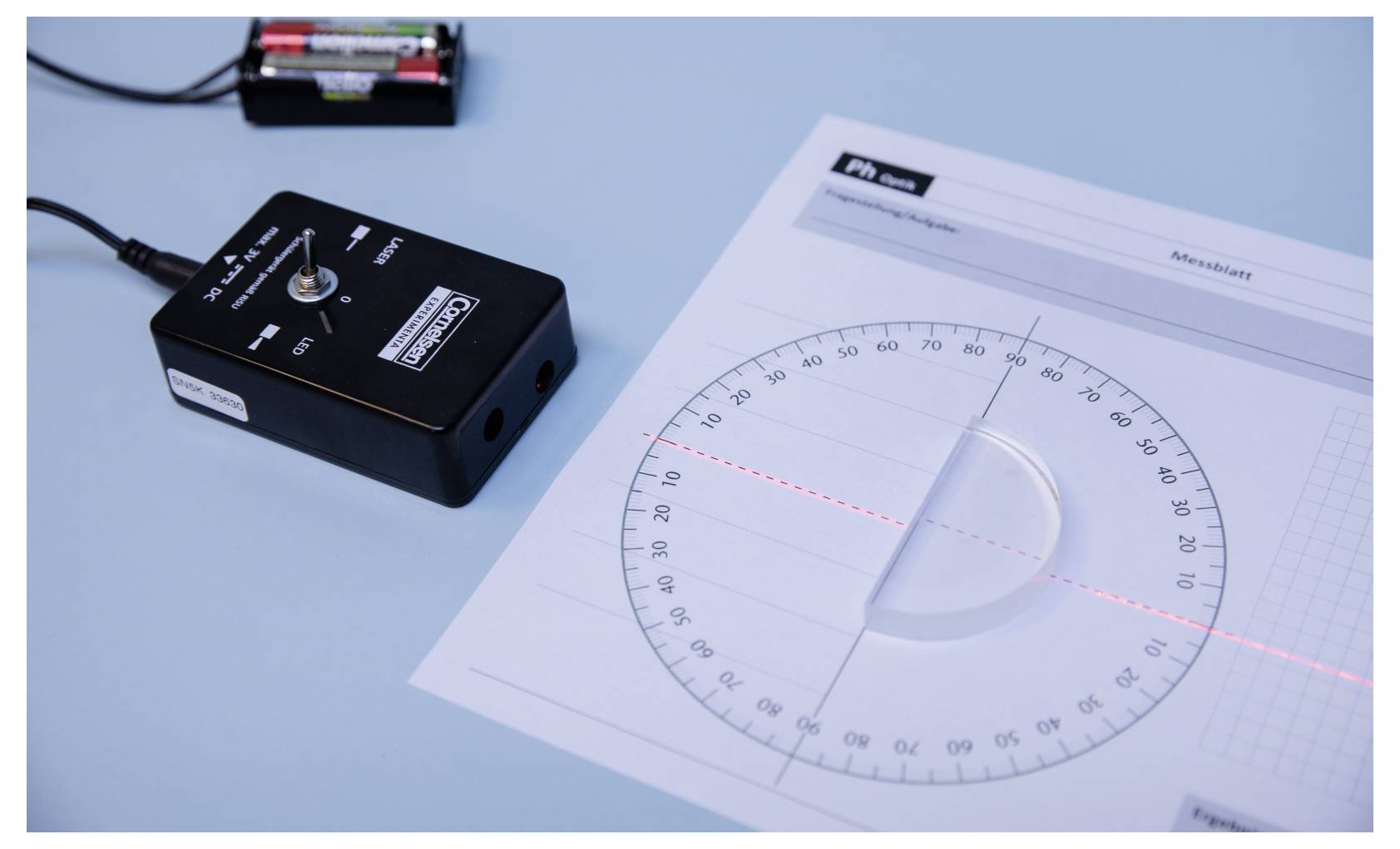 A protractor with a laser beam intersects the center, highlighting angles. Nearby, a power unit with connected wires is visible on a blue surface.