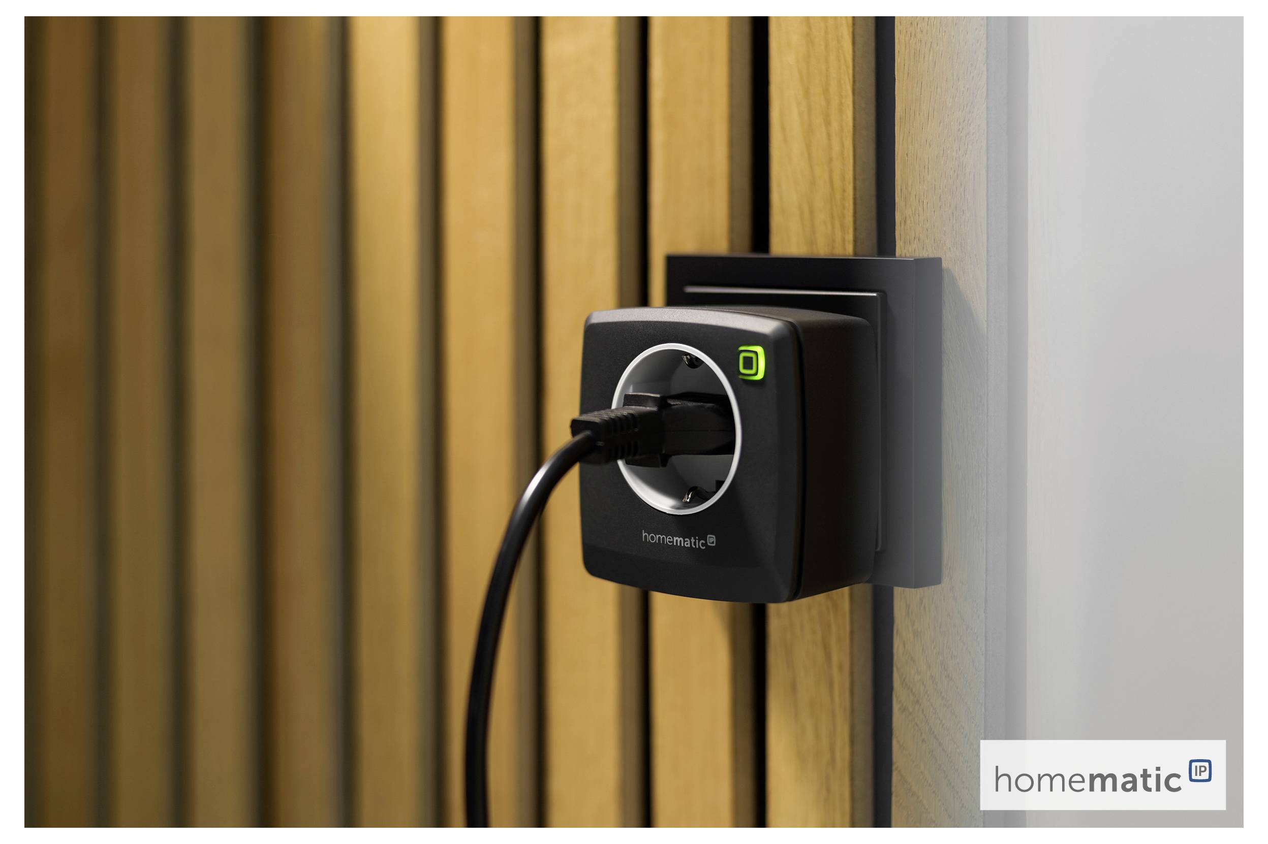A smart plug installed on a wooden wall with a device plugged into it; a logo 'homematic IP' is visible on the plug.