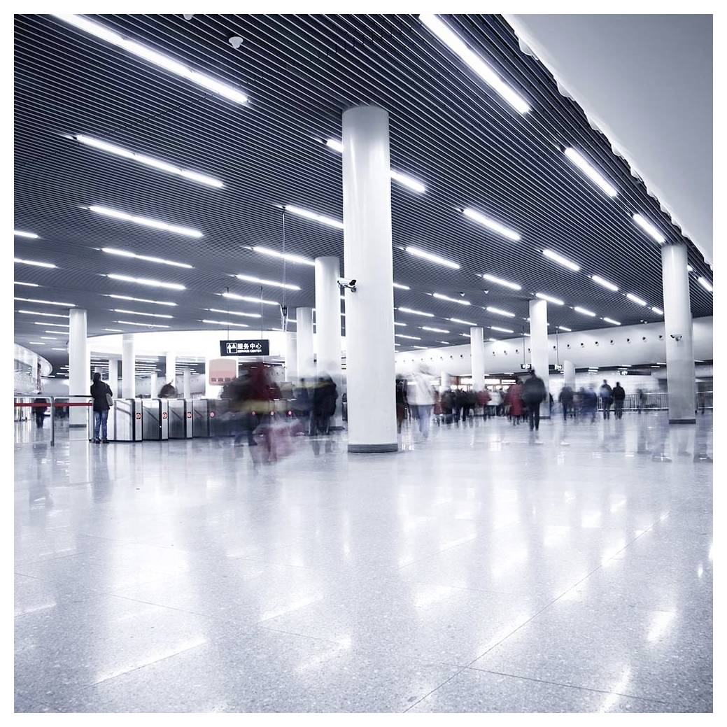 A busy, wide underground transit station with high ceilings and fluorescent lighting, where blurred commuters move through gates.