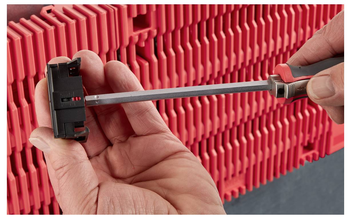 A person uses a screwdriver to adjust a black component against a red, ribbed background, likely part of a mechanical or electrical setup.