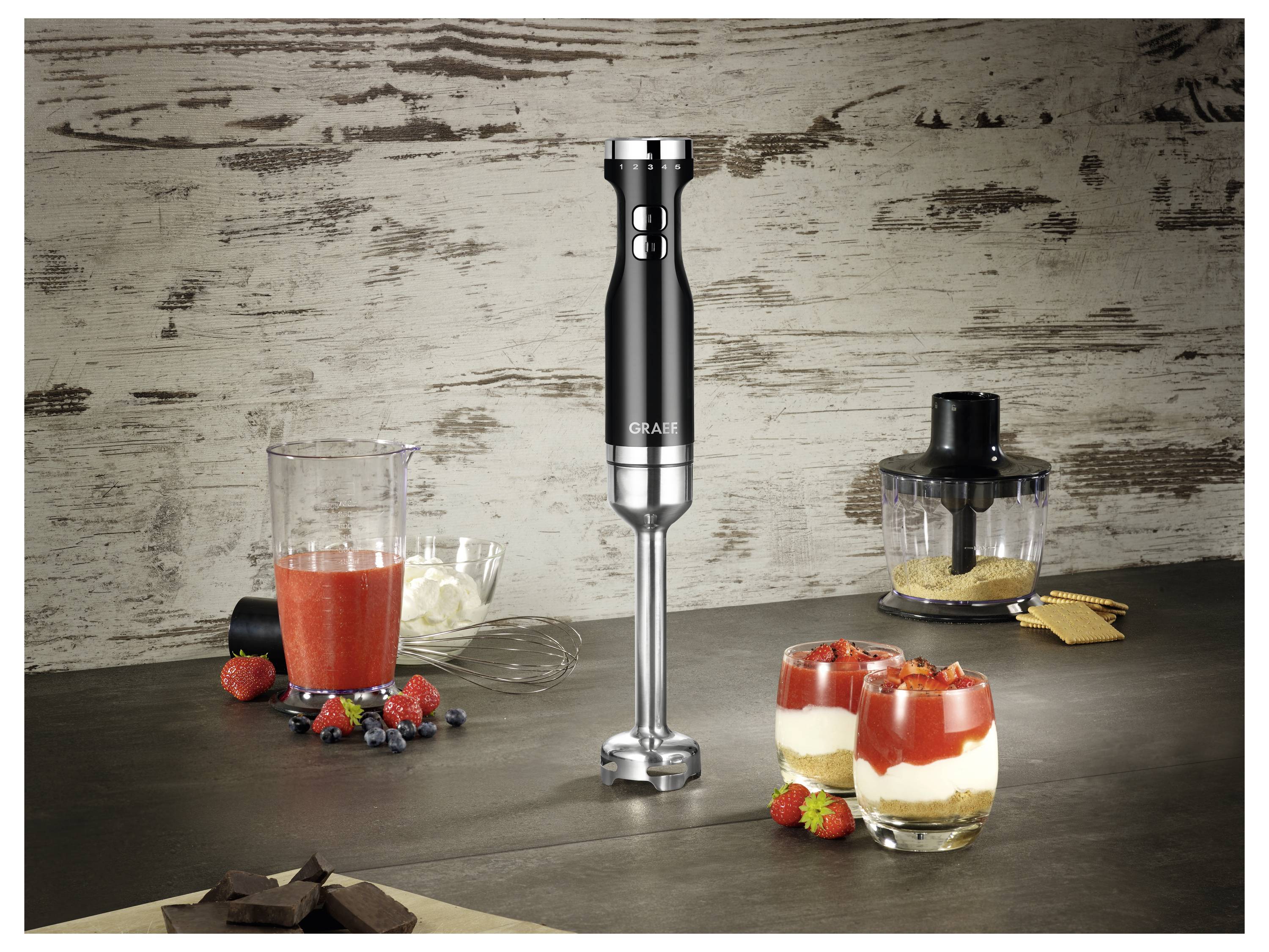 A black immersion blender on a kitchen counter next to glasses of layered dessert, fresh berries, chocolate, and kitchenware.