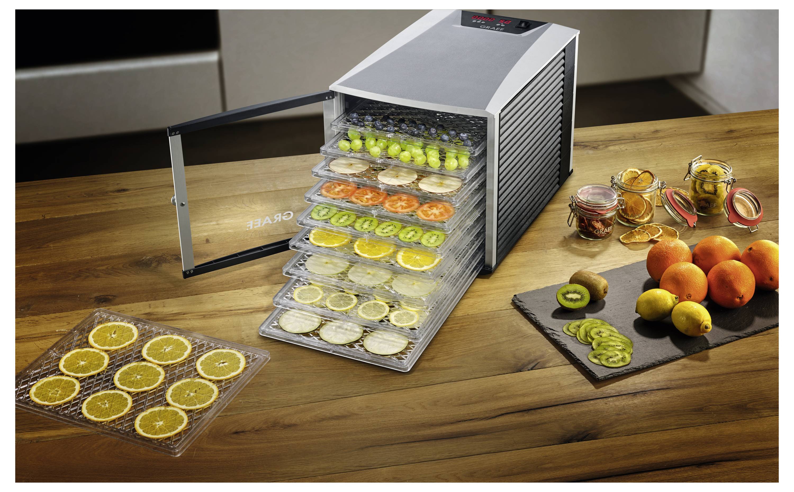 A food dehydrator with trays of sliced fruits and vegetables. Nearby, fresh oranges and kiwis are placed on a wooden surface.