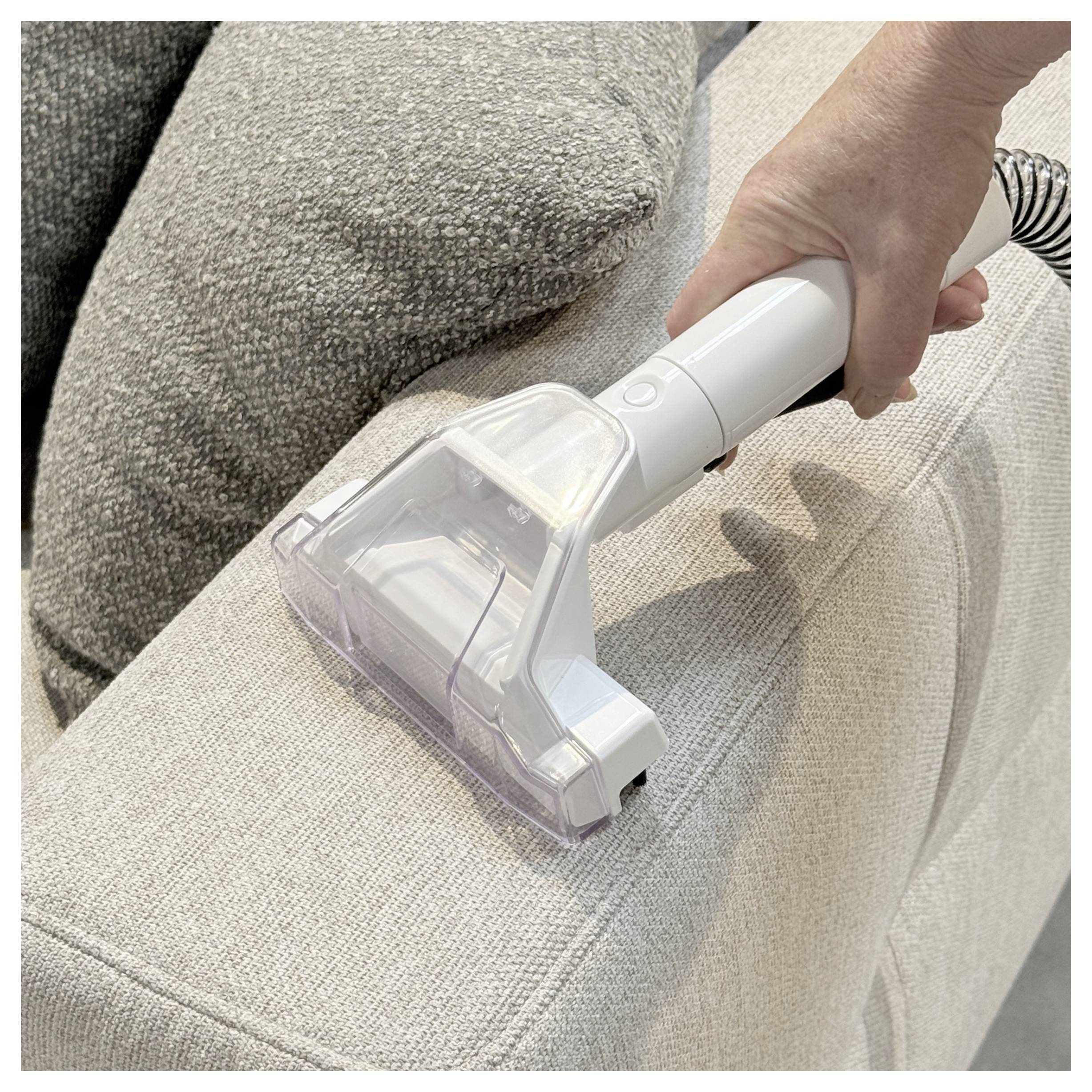 A person uses a handheld vacuum on a light-colored upholstered couch, focusing on cleanliness and maintenance.