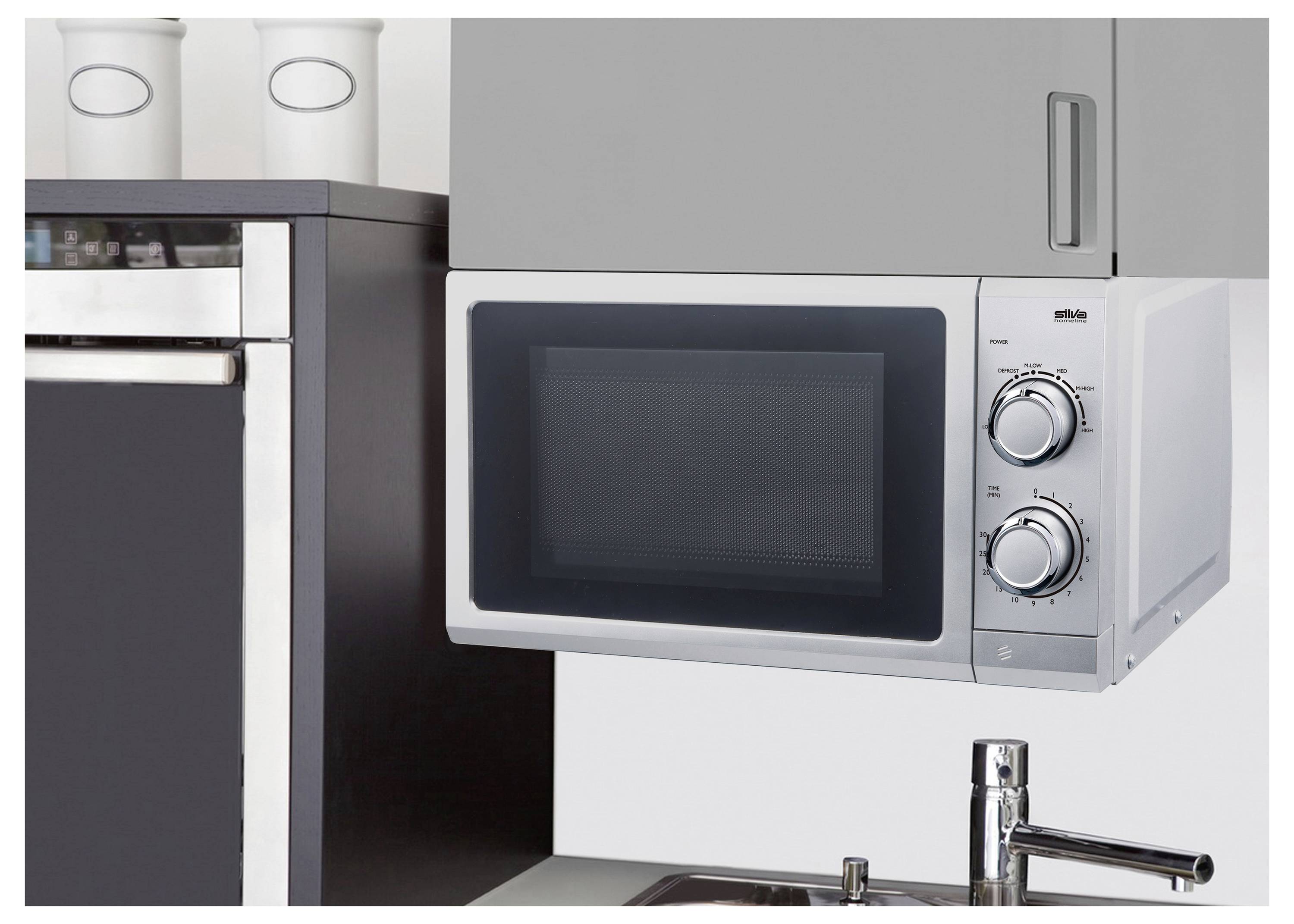 A wall-mounted microwave oven with a black and silver finish, featuring two dials for settings. It is positioned above a sink and next to a cupboard.