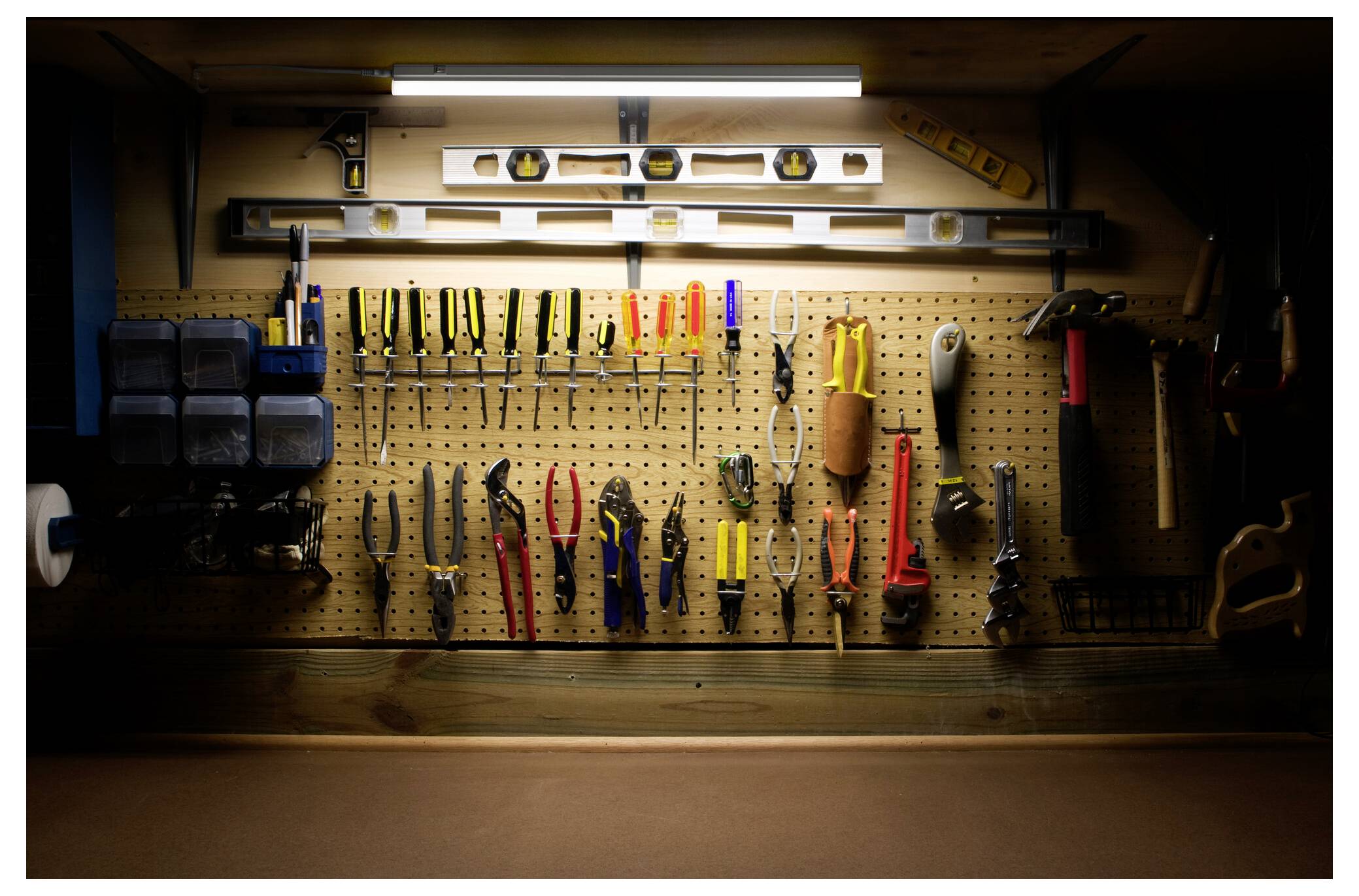 A pegboard in a dimly lit workshop displays organized hand tools, including pliers, screwdrivers, and hammers, with levels on top.