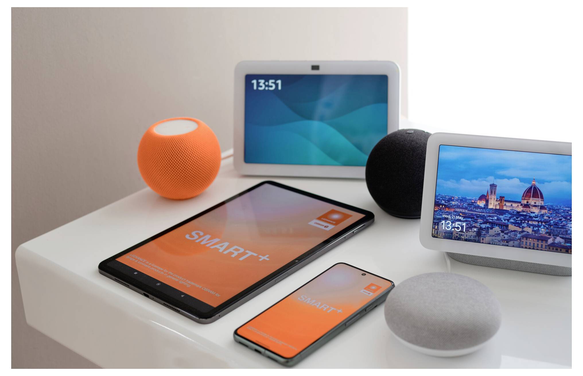 Smart home devices on a table: tablet, smartphone, smart speakers, and displays, showing time and scenic backgrounds.
