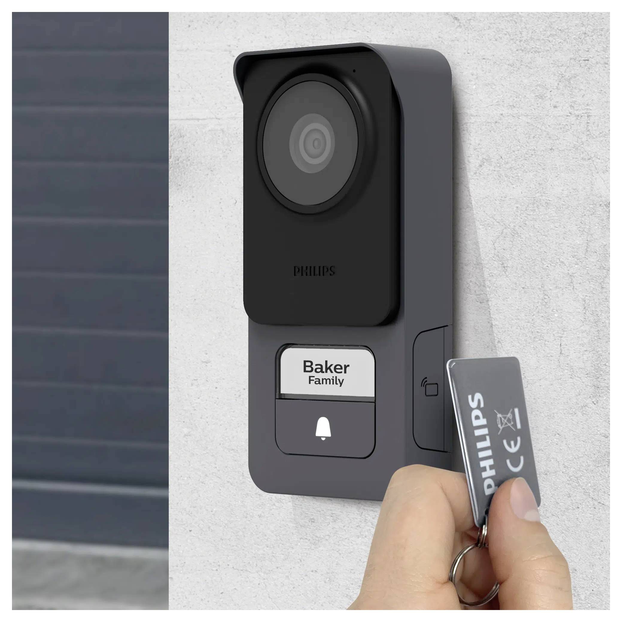 A hand holds a 'Philips' key card near a black, wall-mounted video doorbell with a 'Baker Family' name tag and a doorbell button.
