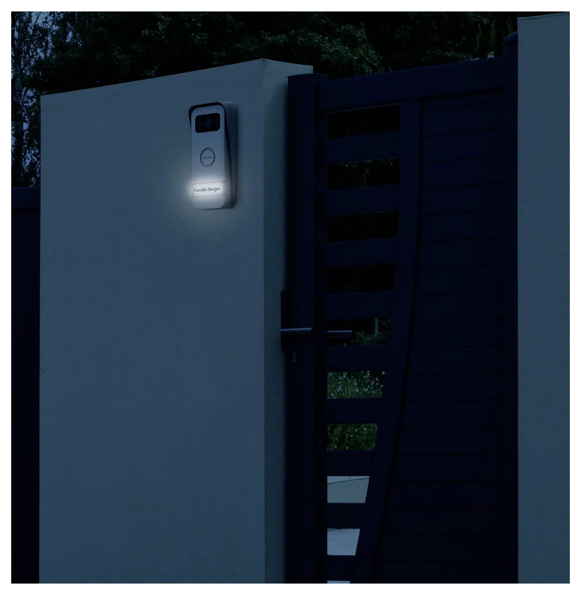 A modern doorbell mounted on a wall next to a closed gate glows softly in the dark, indicating its location and readiness.