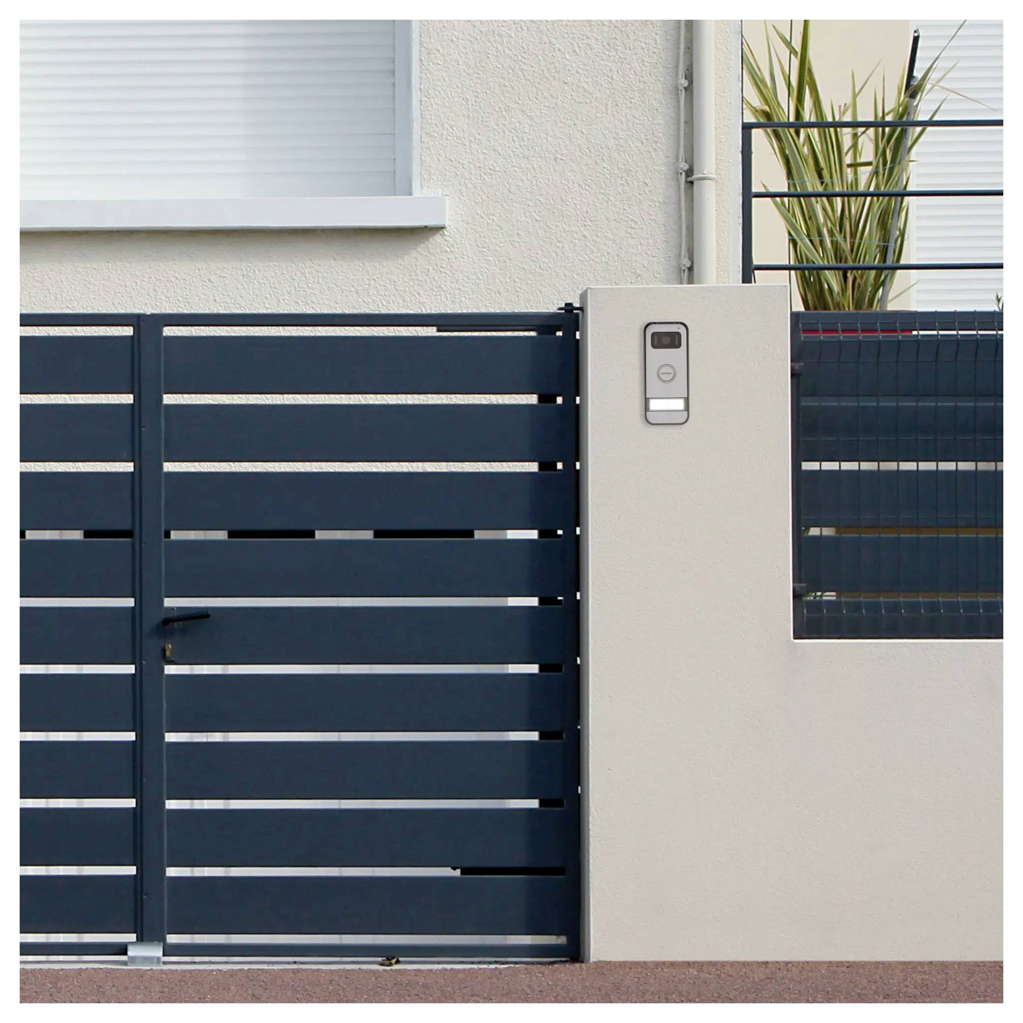 Modern black metal gate and intercom on a concrete pillar in front of a white building, with a potted plant on the balcony above.