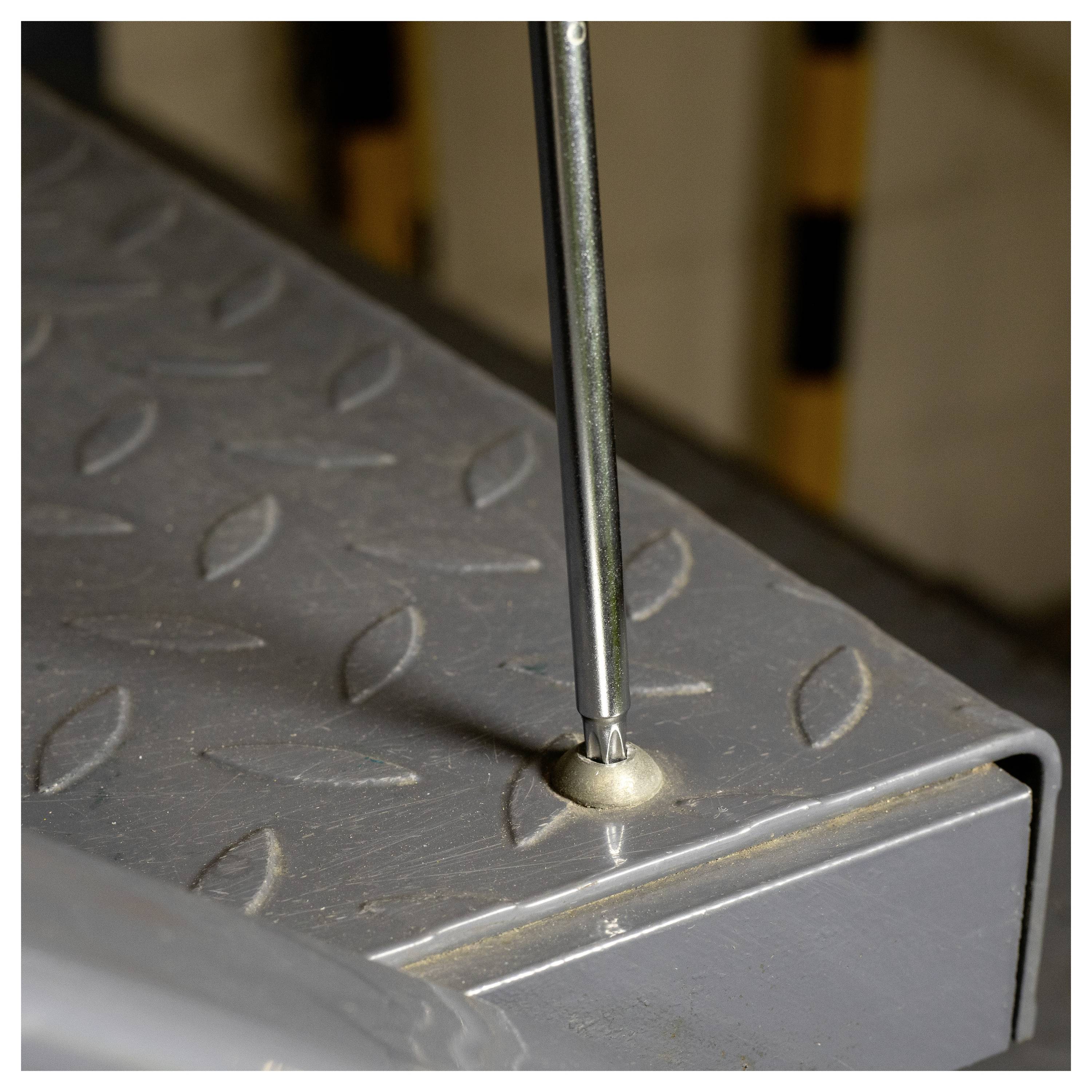 A screwdriver is inserted into a screw on a metal surface with diamond plate texture. The background shows yellow and black striped markings.