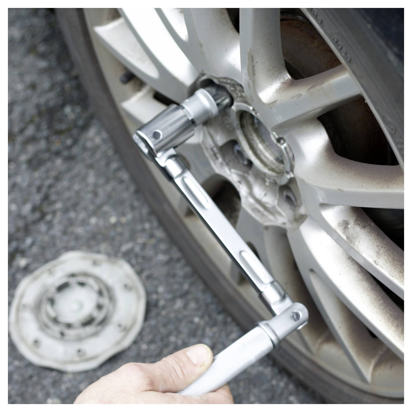 A hand uses a lug wrench to remove a bolt from a car wheel, with a removed hubcap lying on the ground nearby.
