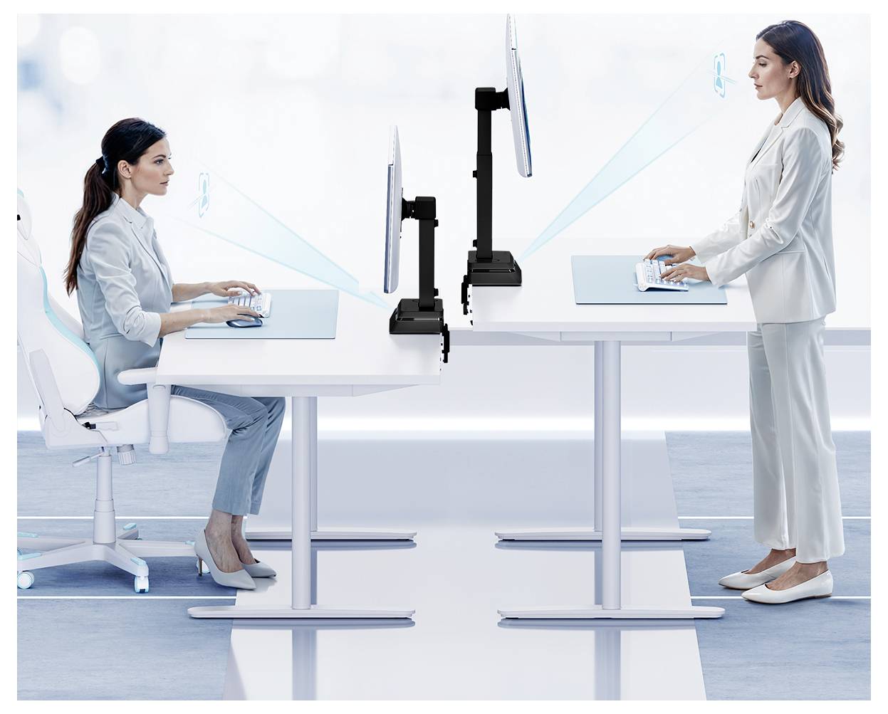 Two women in professional attire work at desks facing each other, one sitting and one standing, both using computers with screen arms.