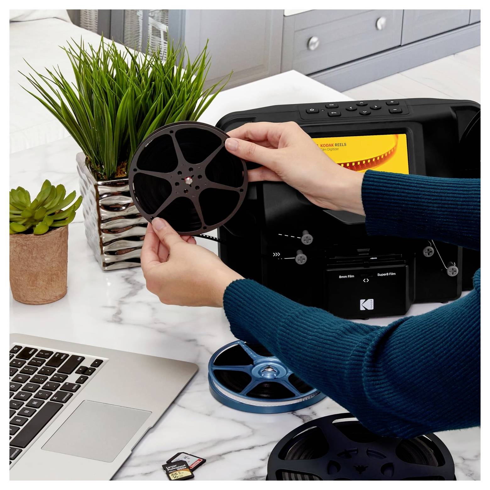 Hands hold a film reel above a scanning device, with a laptop and memory cards nearby on a marble surface.