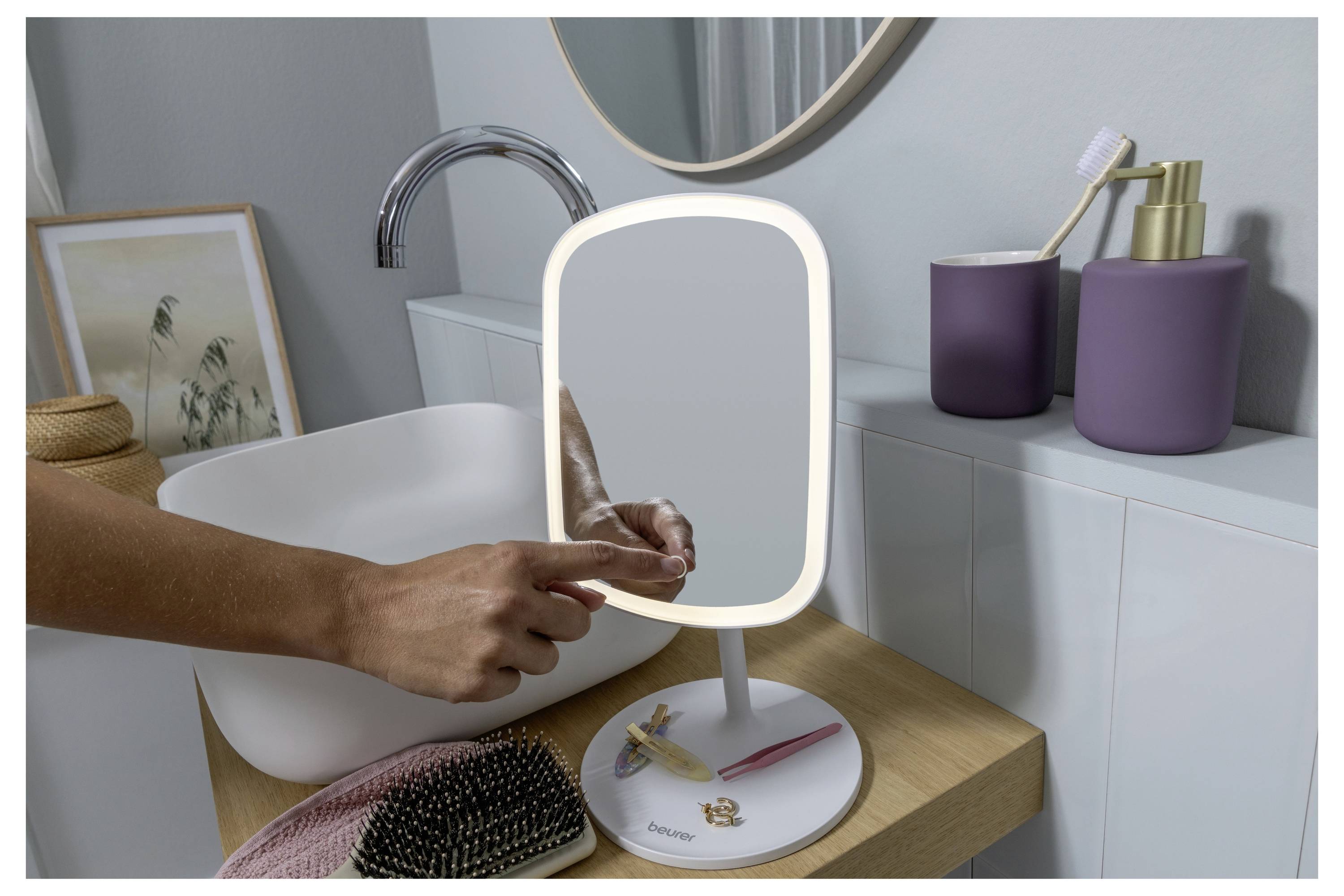 A person is touching a tabletop mirror with a glowing frame, set on a wooden bathroom counter with a sink, painting, and toiletries.