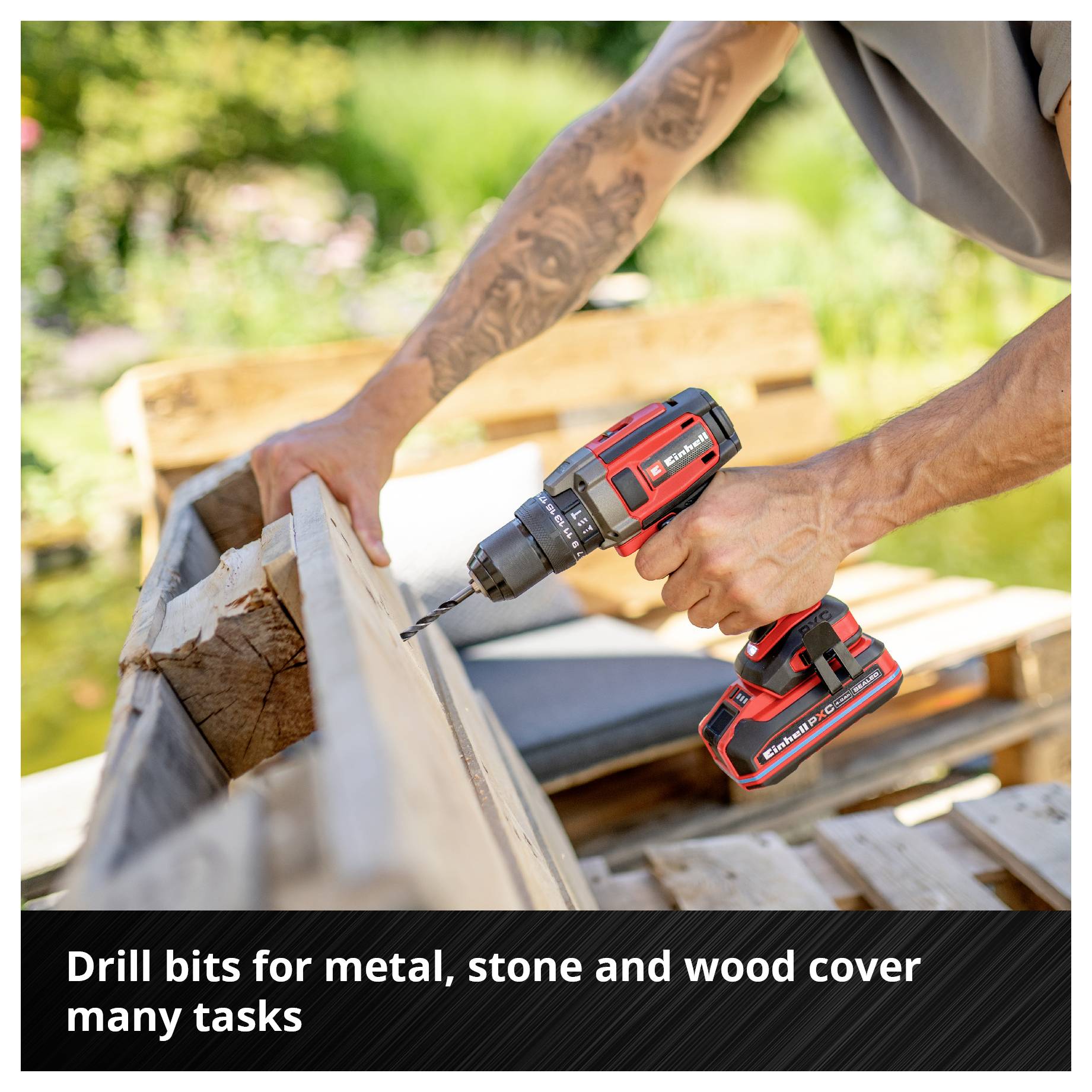 A red cordless drill is being used by a person to drill wood. Blurred garden furniture can be seen in the background.